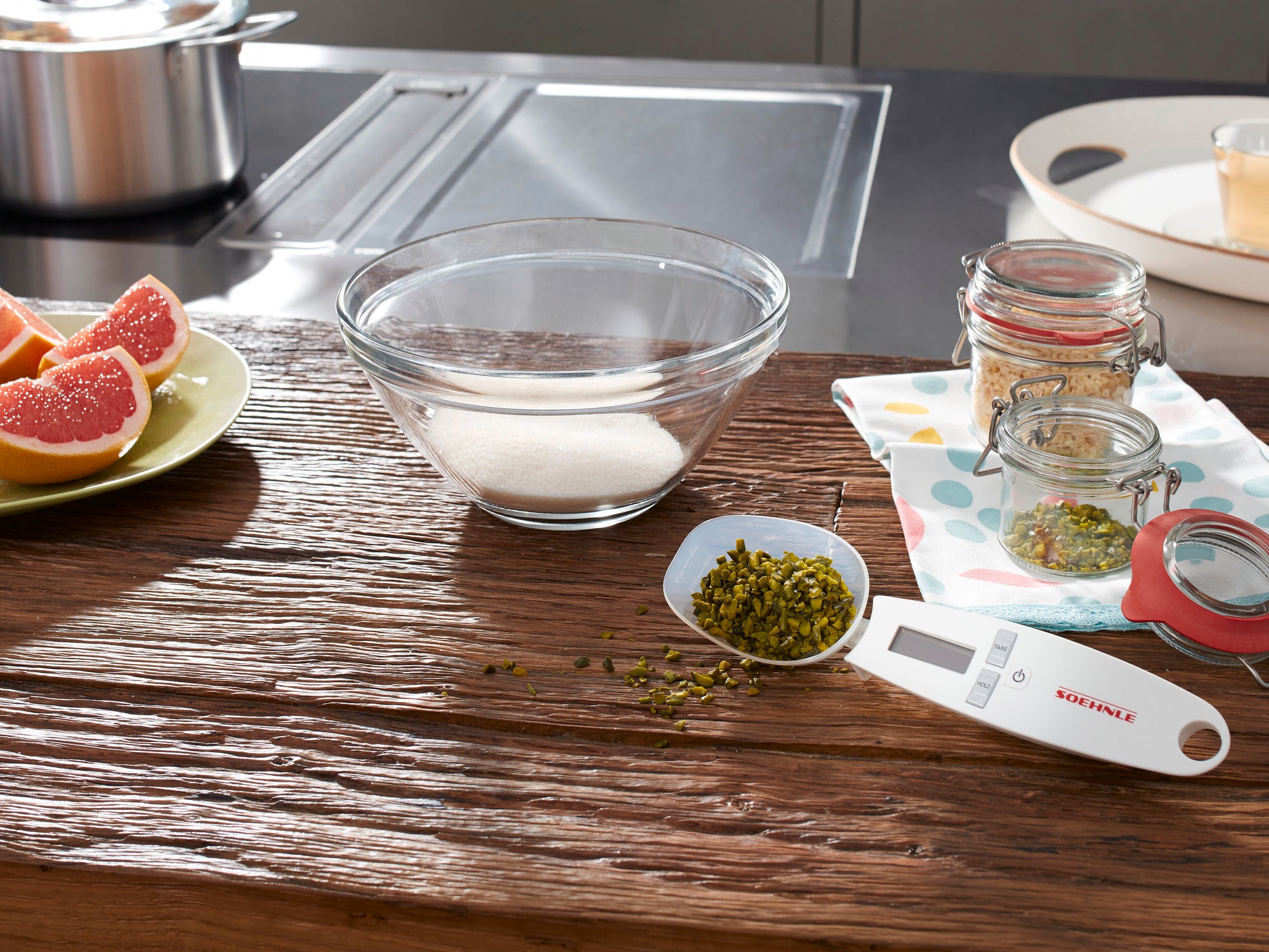 Cyfrowa waga kuchenna Cooking Star leży na rustykalnym drewnianym stole. Obok znajduje się miska z cukrem, słoiki z przyprawami i świeży grejpfrut.