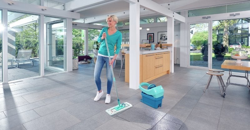 Eine Frau wischt mit einem Mopp den grossen, modernen und hellen Wohnbereich mit offenen Fenstern und einer Holz-Glas-Kueche.