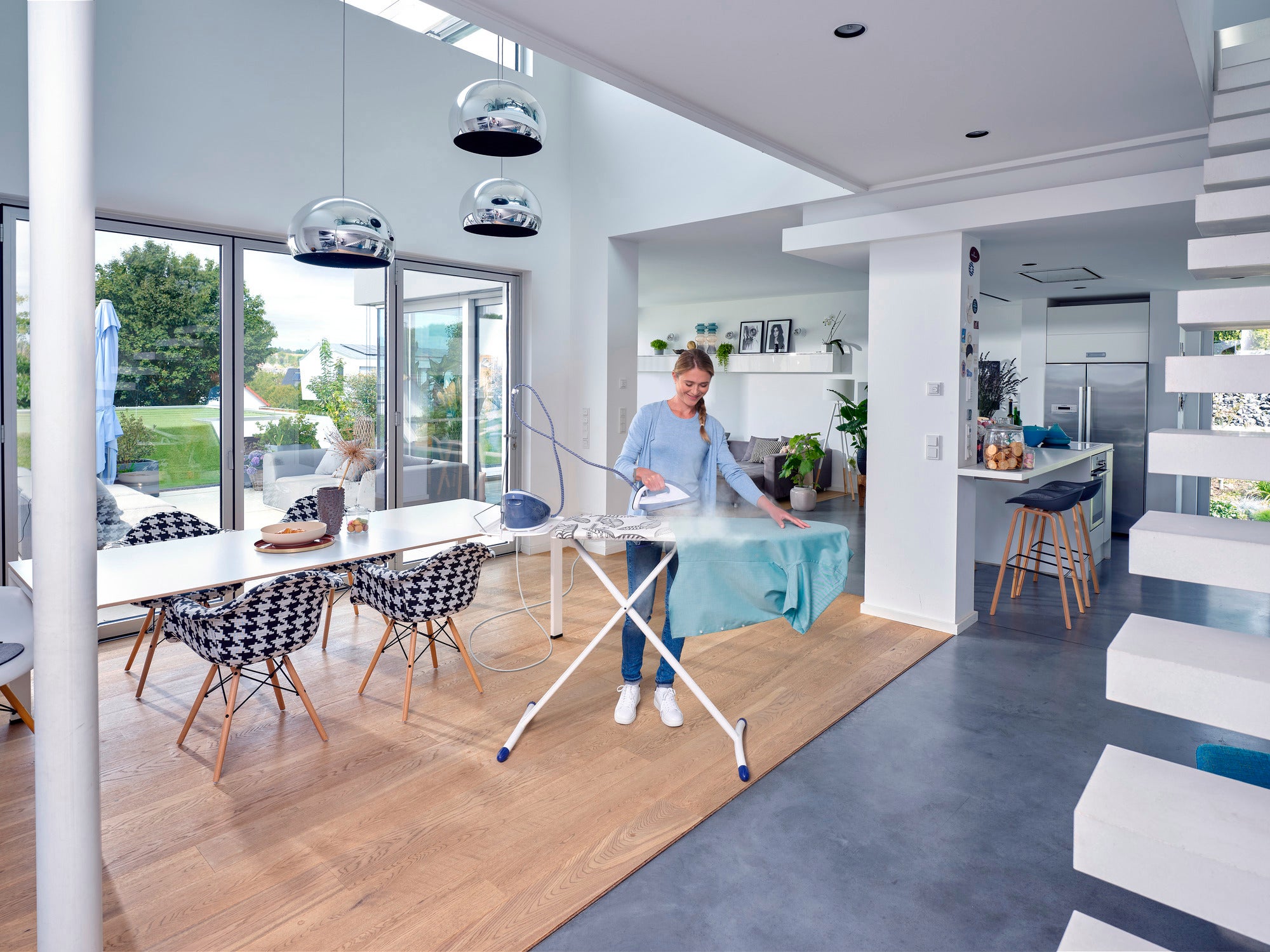 Une personne repasse des vêtements sur la table à repasser Air Board Express M Solid dans une pièce moderne et lumineuse avec de grandes fenêtres et un mobilier élégant. Idéal pour un repassage efficace.