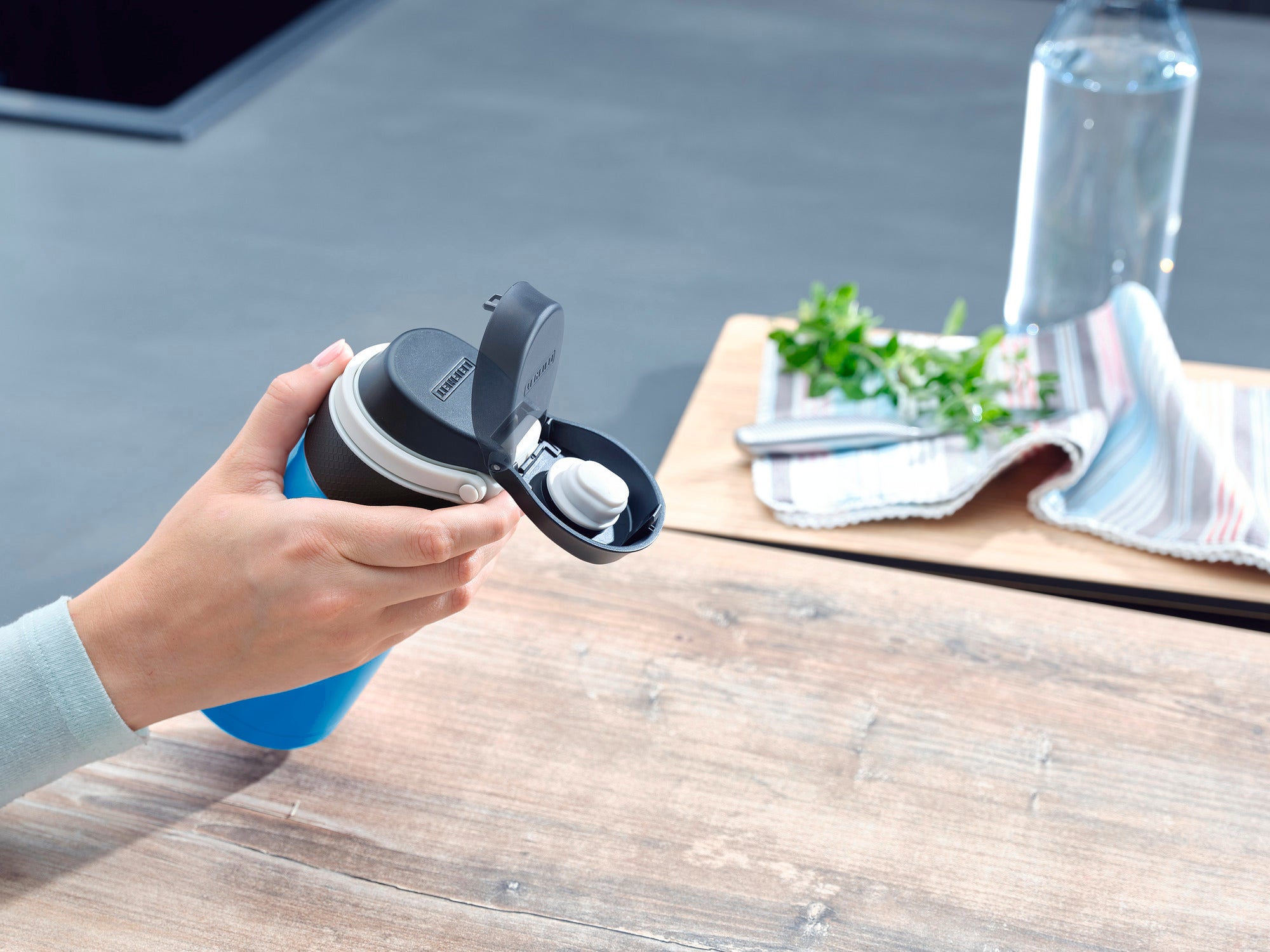 Eén persoon houdt een lichtblauwe geïsoleerde mok Flip vast met een inhoud van 350 ml. De mok heeft een praktisch klapdeksel. Op de achtergrond zijn een tafel, een handdoek en een waterfles te zien.