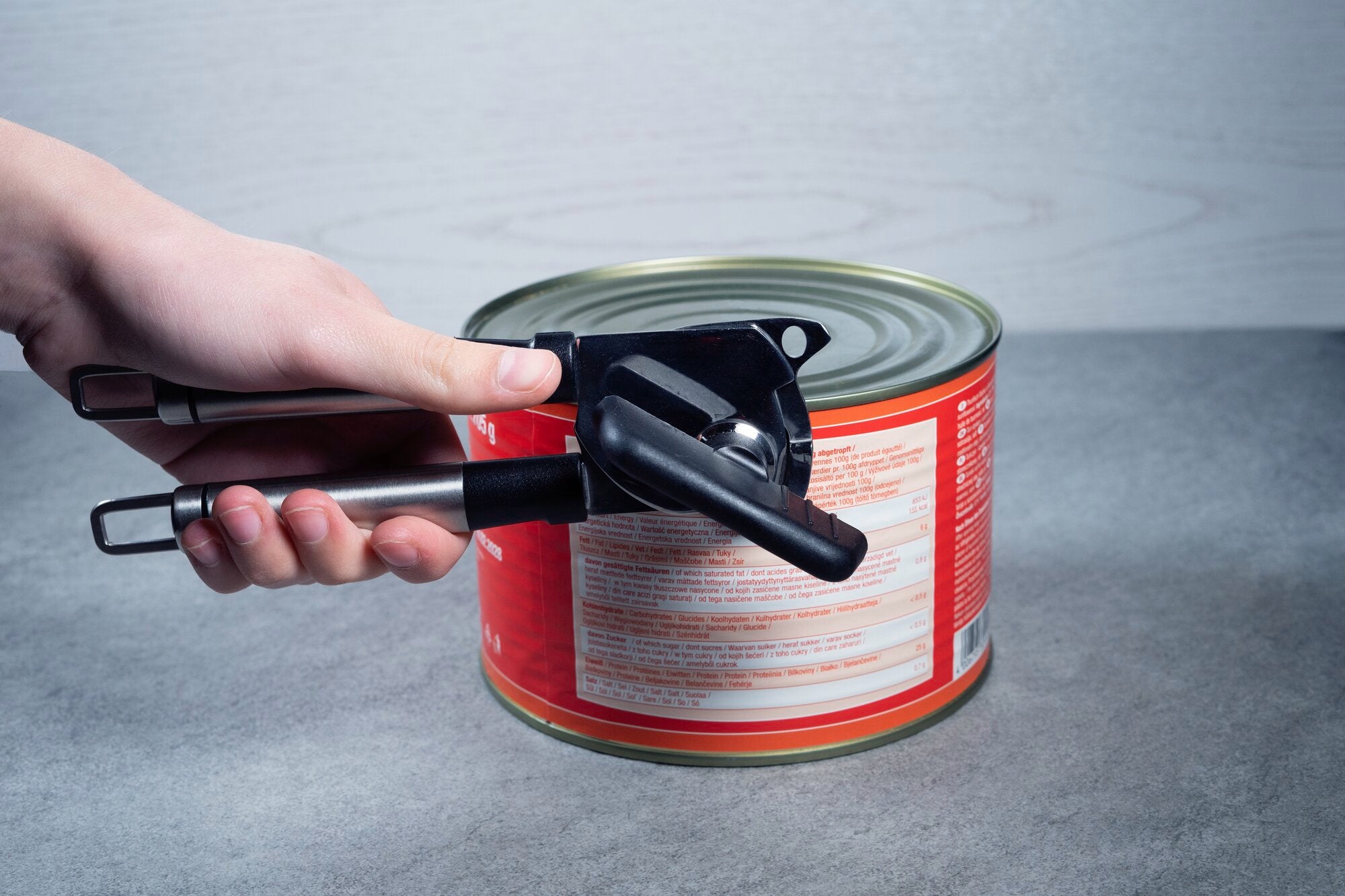 Een hand gebruikt een metalen blikopener om een groot, rood blik met voedsel op een grijze oppervlak te openen.