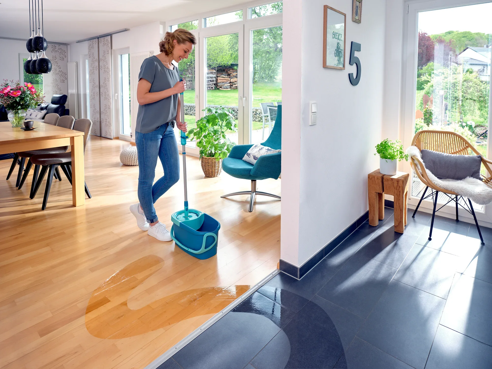 Una donna pulisce il pavimento in legno di un soggiorno moderno con il lavapavimenti CLEAN TWIST Ergo. La stanza è luminosa e arredata con stile, con grandi finestre e un tavolo da pranzo. Il lavapavimenti è ideale per la pulizia accurata di superfici del