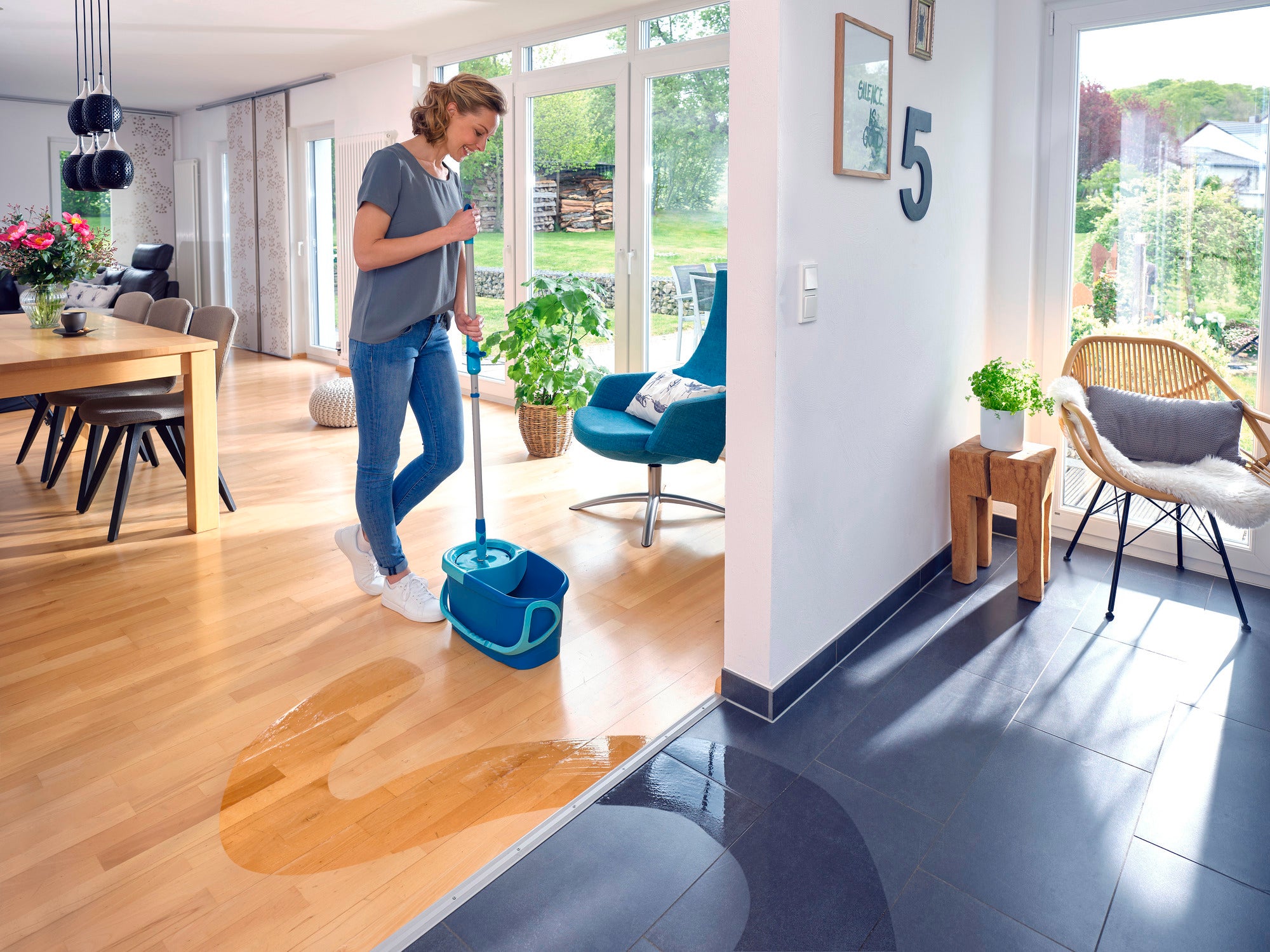 Kobieta czyści drewnianą podłogę w nowoczesnym salonie za pomocą CLEAN TWIST Mop Ergo. Pokój jest jasny i stylowo urządzony, z dużymi oknami i stołem jadalnym. Mop jest idealny do dokładnego czyszczenia delikatnych powierzchni.
