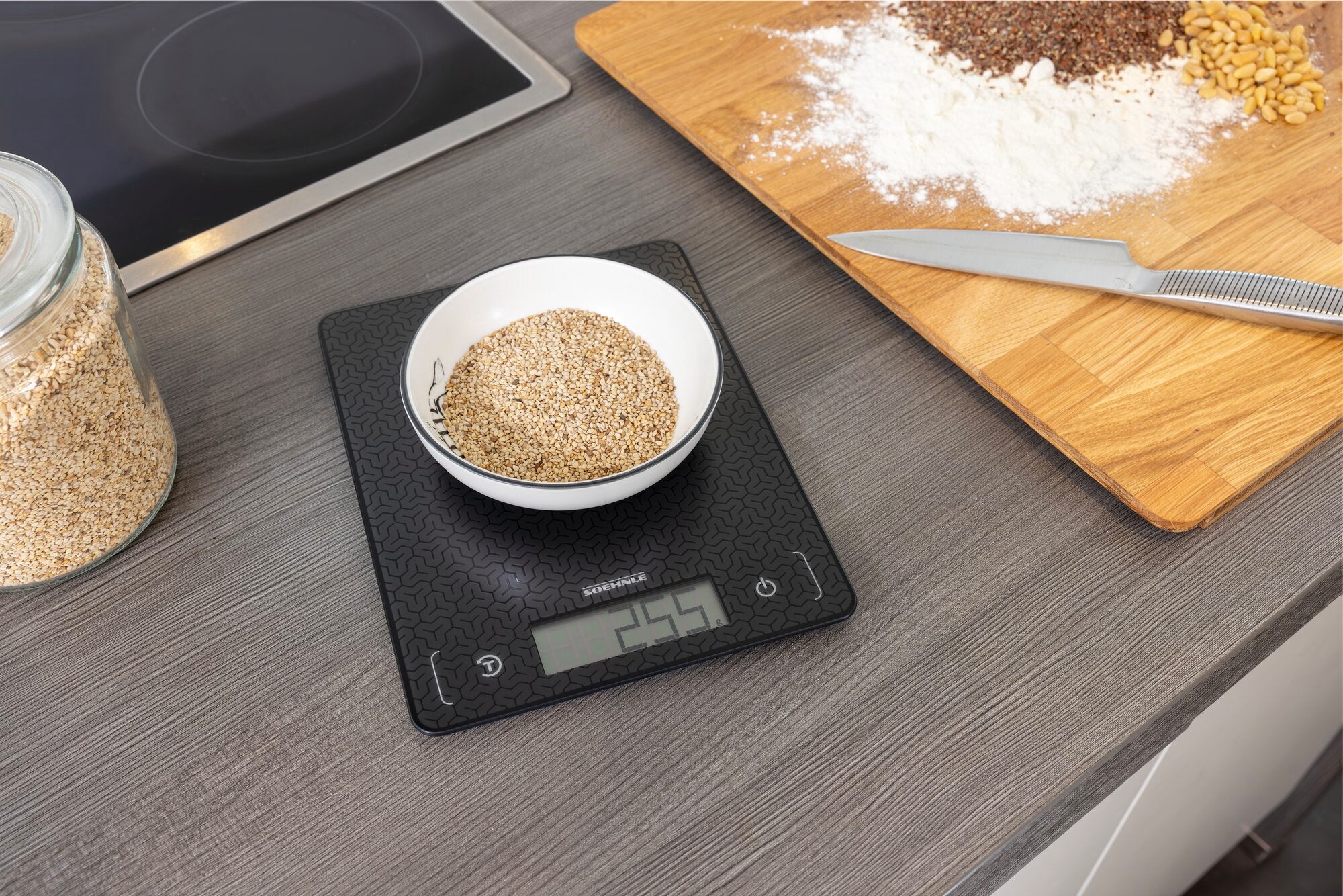 Digitale Küchenwaage Comfort Mosaic auf einer Arbeitsplatte. Eine Schüssel mit Getreide wird gewogen, daneben liegen Mehl und ein Messer auf einem Holzbrett. Ideal für präzises Kochen.