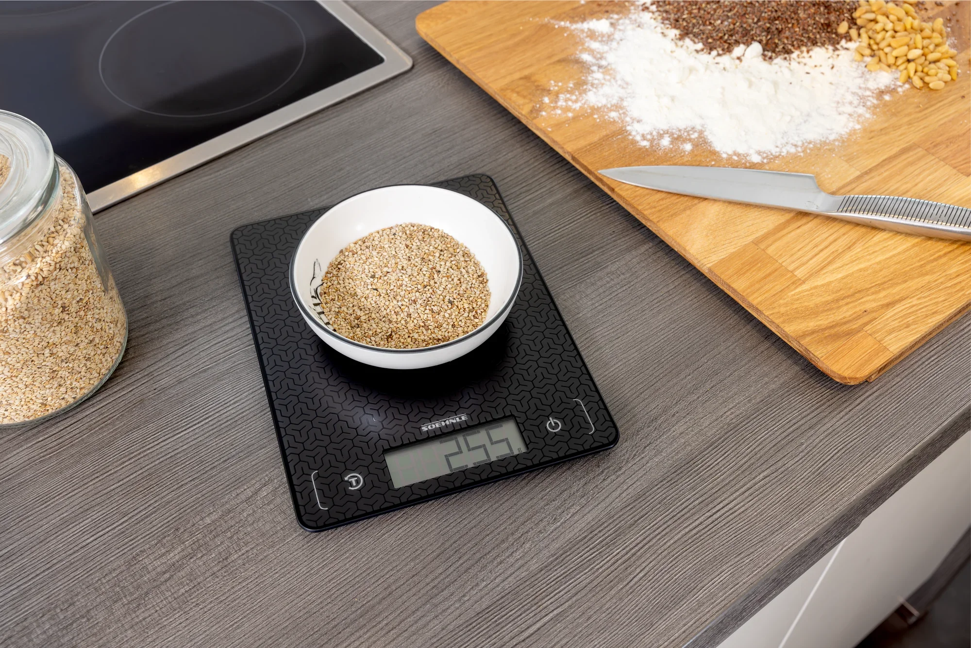 Cyfrowa waga kuchenna Comfort Mosaic na blacie kuchennym. Miska z ziarnem jest ważona, a obok na drewnianej desce znajduje się mąka i nóż. Idealna do precyzyjnego gotowania.