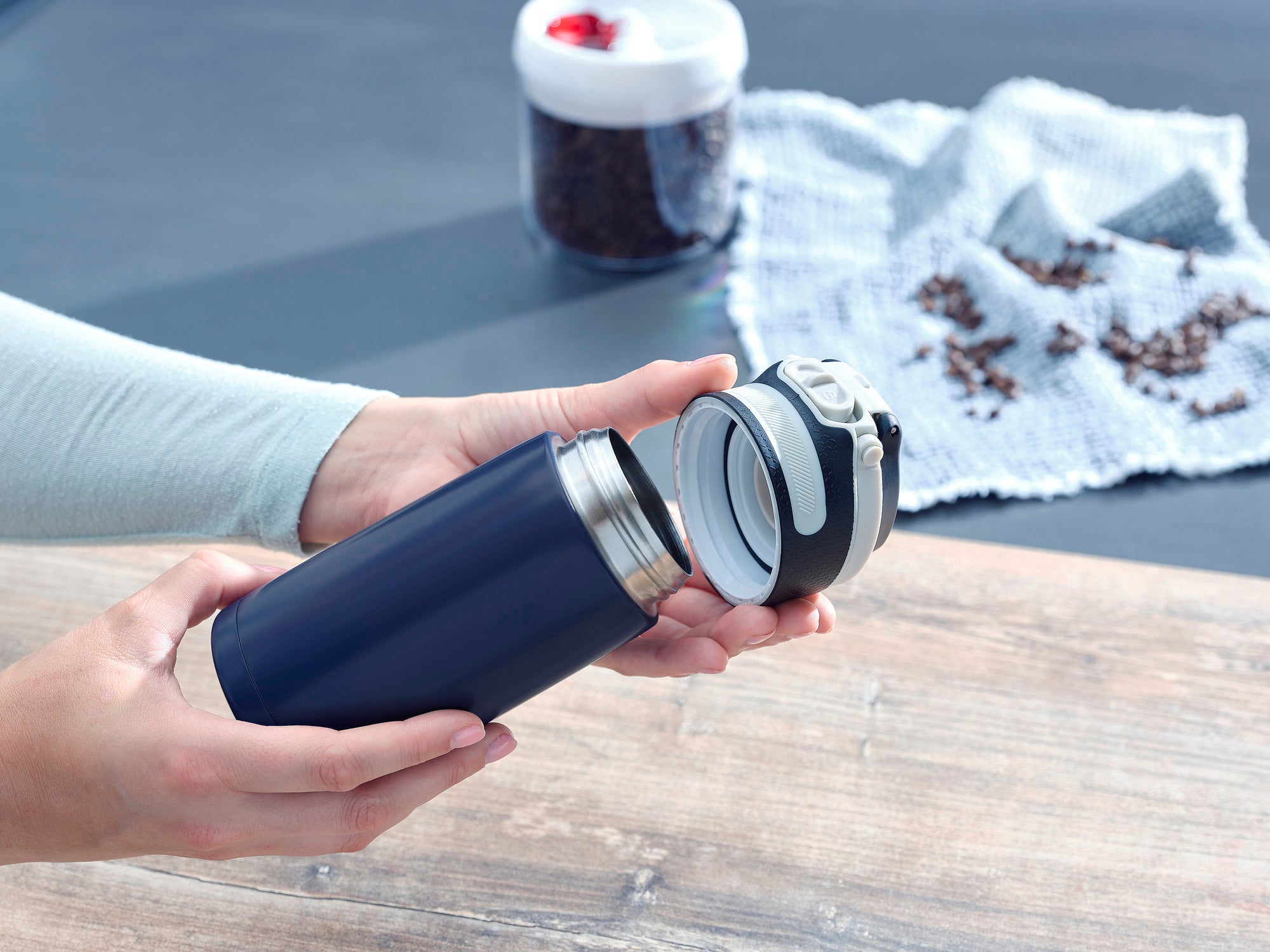 Isolierbecher Flip 350ml in Dunkelblau wird von einer Person geöffnet. Im Hintergrund sind Kaffeebohnen und ein weißes Tuch zu sehen. Ideal für unterwegs.