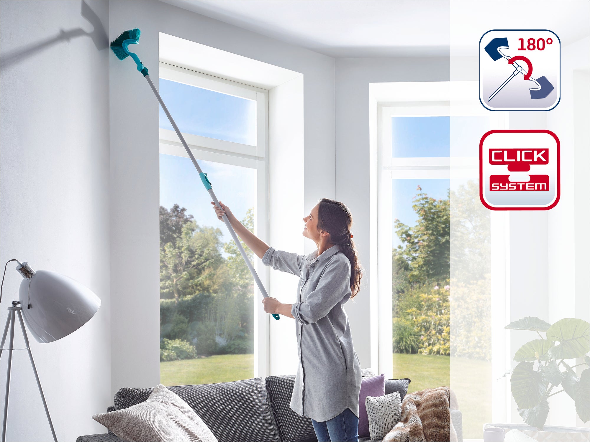 A woman cleans a high window corner with a Leifheit window squeegee with a telescopic handle. The squeegee has a 180-degree joint. Ideal for hard-to-reach places.