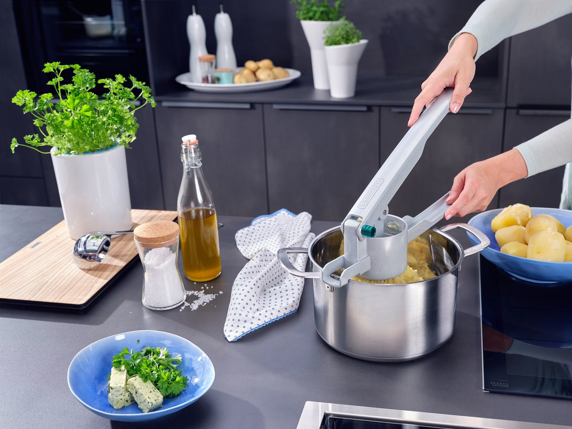 Une personne utilise un presse-pommes de terre pour presser des pommes de terre cuites dans une casserole. Des épices, de l'huile et des herbes fraîches sont disposées sur le plan de travail.