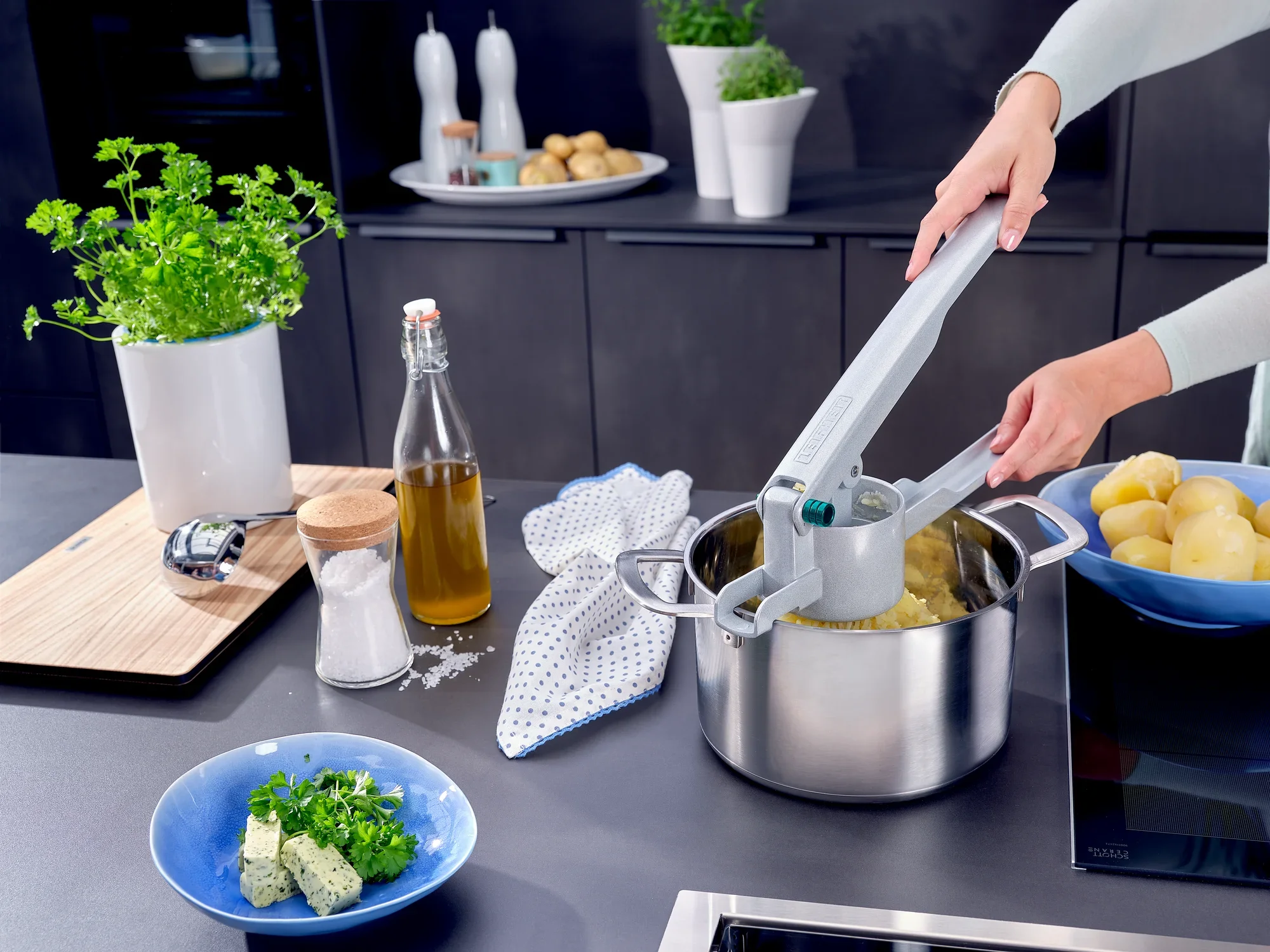 Jedna osoba używa praski do ziemniaków spaetzle do wyciskania ugotowanych ziemniaków w garnku. Przyprawy, olej i świeże zioła są gotowe na powierzchni roboczej.