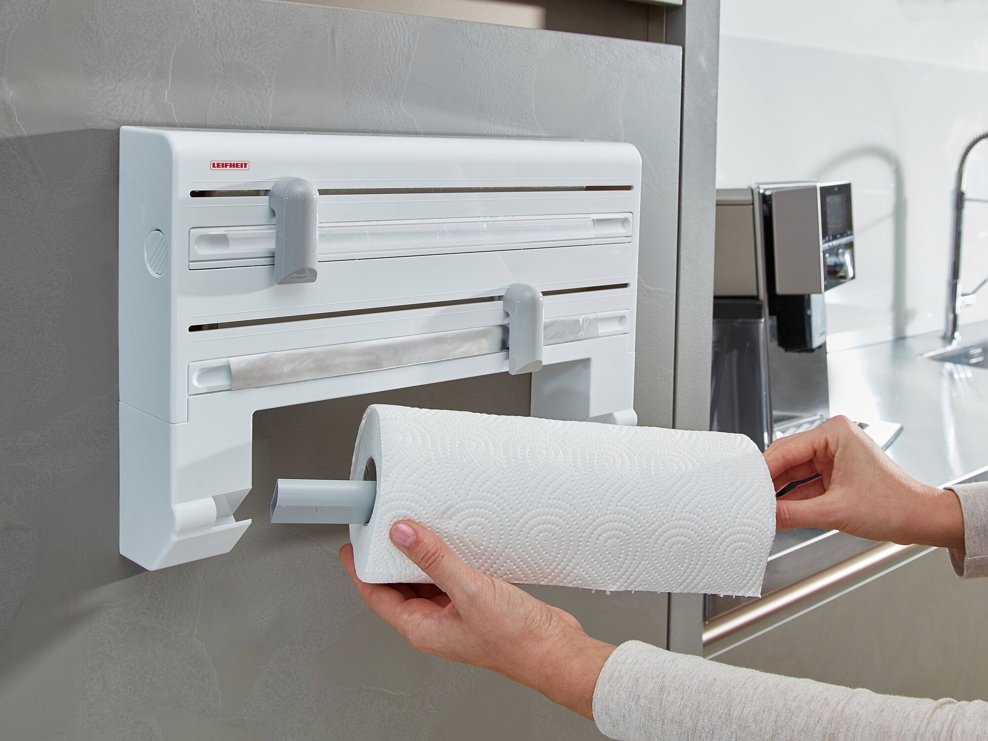 Support mural pour rouleaux Parat : une personne installe un rouleau de papier essuie-tout dans un support mural blanc. Le support est fixé à un mur de cuisine moderne et offre un accès facile.