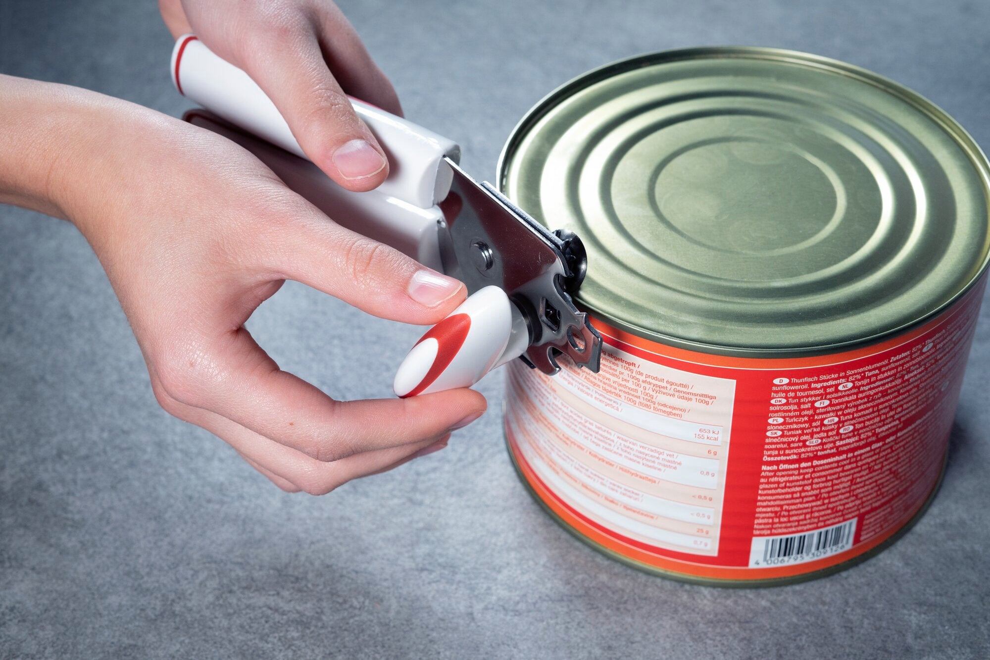 Mani che aprono una lattina di metallo con un apriscatole manuale bianco e rosso, su una superficie grigia.