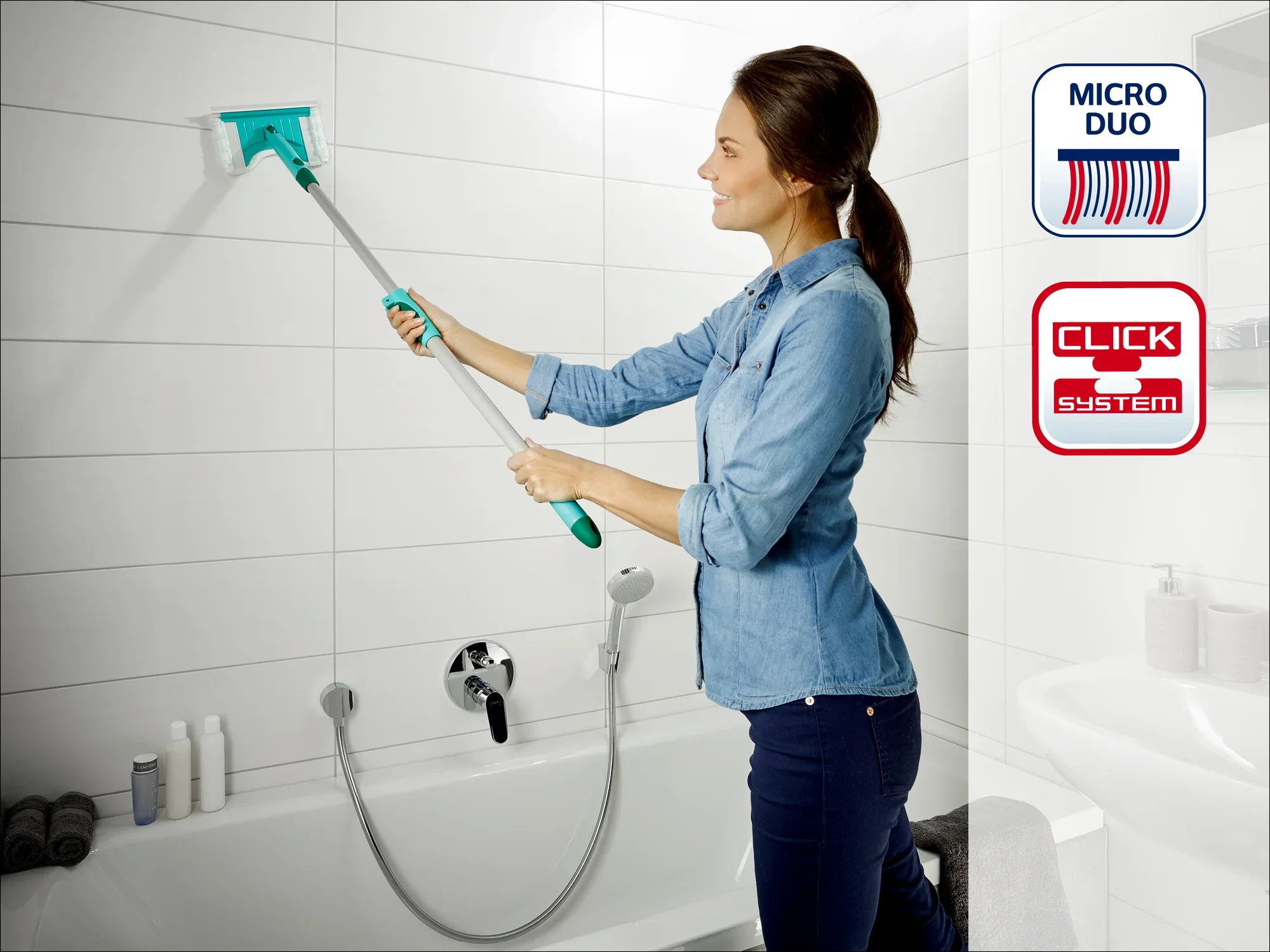 Kobieta czyści płytki łazienkowe za pomocą ściągaczki Leifheit Click System. Ściągaczka ma nakładkę Micro Duo Pad, idealną do dokładnego czyszczenia. W tle widoczne są przybory łazienkowe.