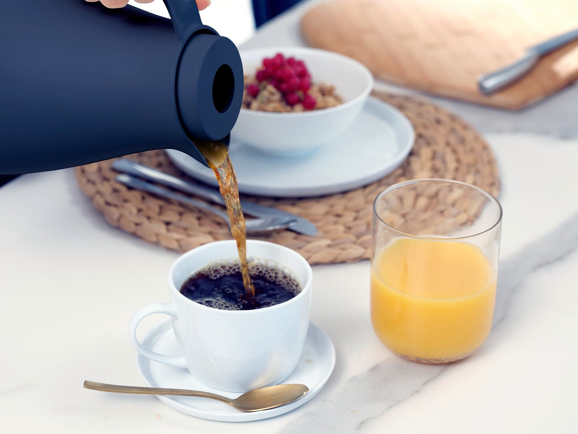 A black vacuum flask called Harmonic pours hot coffee into a white cup. Next to it, a glass of orange juice and a bowl of muesli stand on a table.