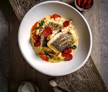 Ein Teller mit gebratenem Fisch auf cremigem Risotto, garniert mit Chorizo, Mandeln und Gemuese, auf einem Holztisch serviert.