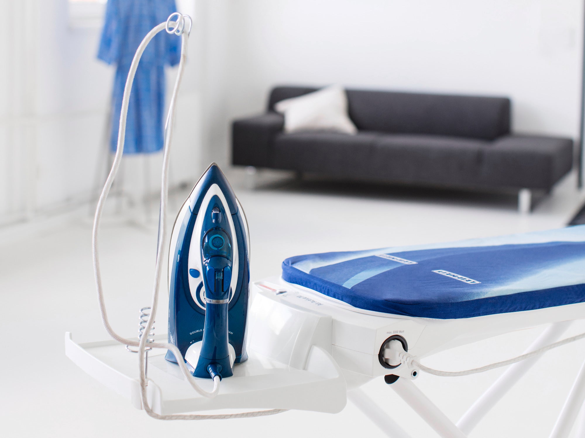 Bügeltisch Air Active M mit einem blauen Bügeleisen auf der Ablage. Im Hintergrund ist ein modernes Wohnzimmer mit einem schwarzen Sofa und einem blauen Bademantel zu sehen.