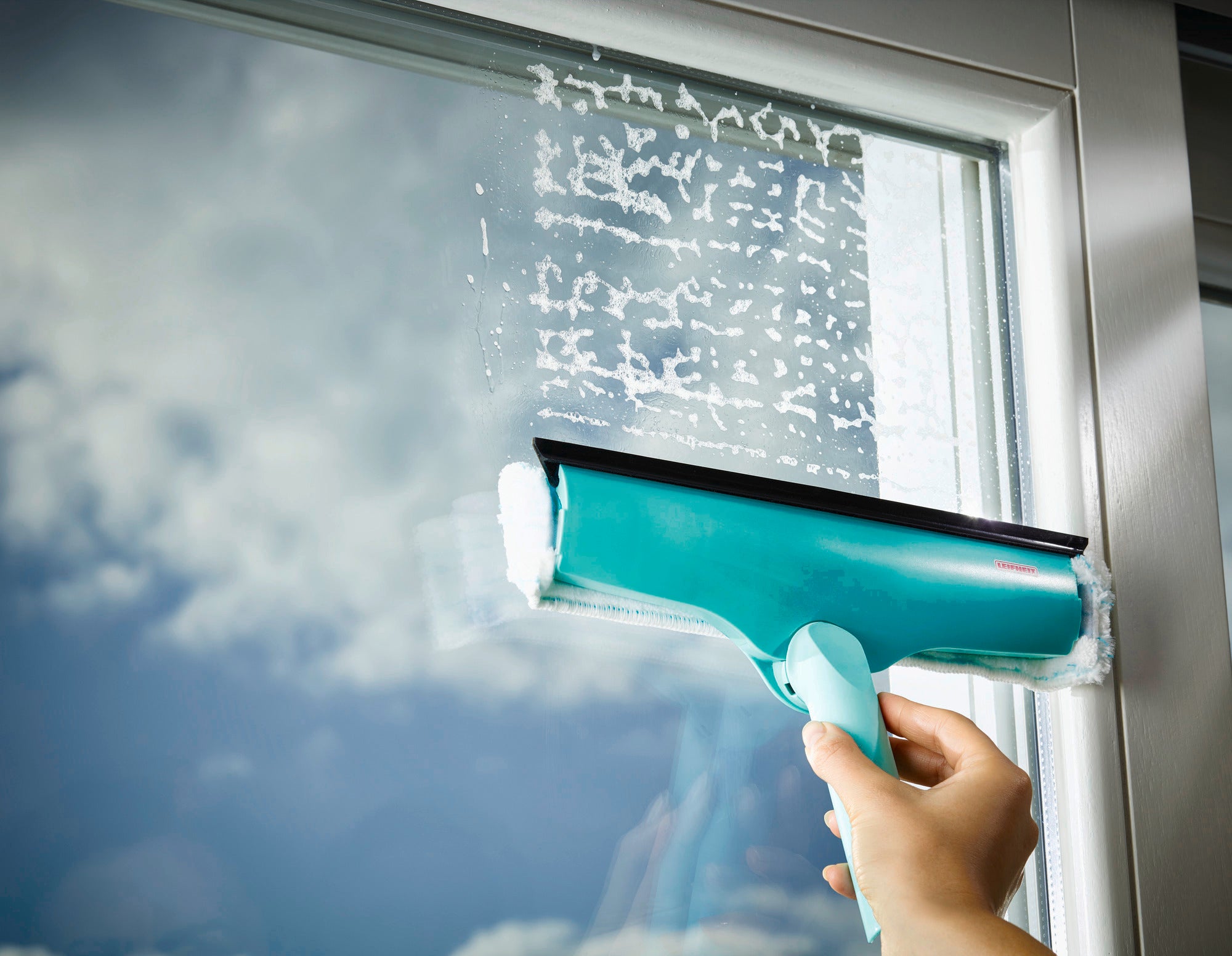 Fensterwischer 3in1 in Aktion: Eine Hand hält den türkisfarbenen Fensterwischer, der über eine Glasfläche gleitet und dabei Schaum entfernt. Der Himmel spiegelt sich im Fenster.