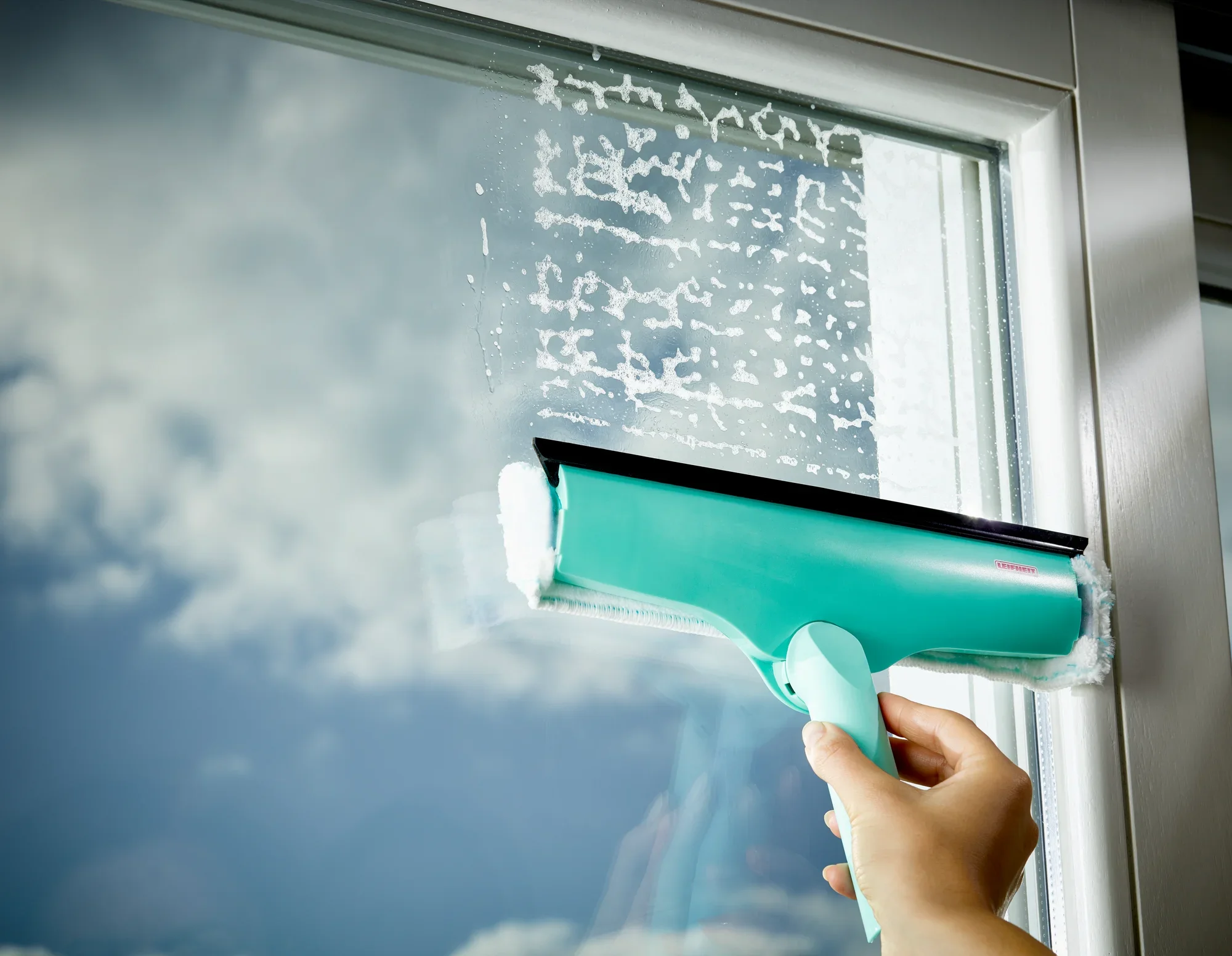 Tergicristallo 3in1 in azione: una mano tiene il tergicristallo turchese che scorre su una superficie di vetro rimuovendo la schiuma. Il cielo si riflette nella finestra.