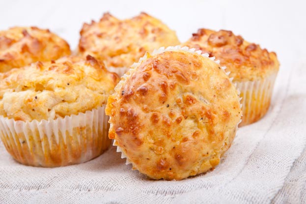 Fuenf goldbraun gebackene Kaesemuffins in Papierfoermchen liegen auf einem weissen Tuch, frisch und koestlich aussehend.