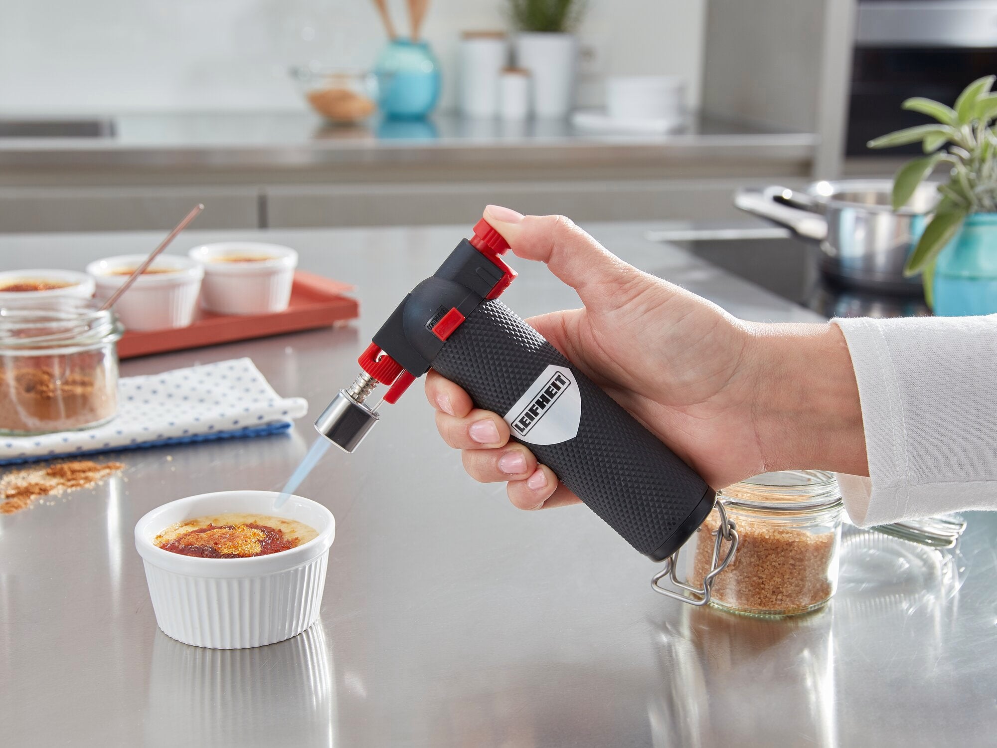 Une main utilise un chalumeau pour carameliser un dessert dans un ramequin blanc sur un plan de travail en inox, avec du sucre a c
