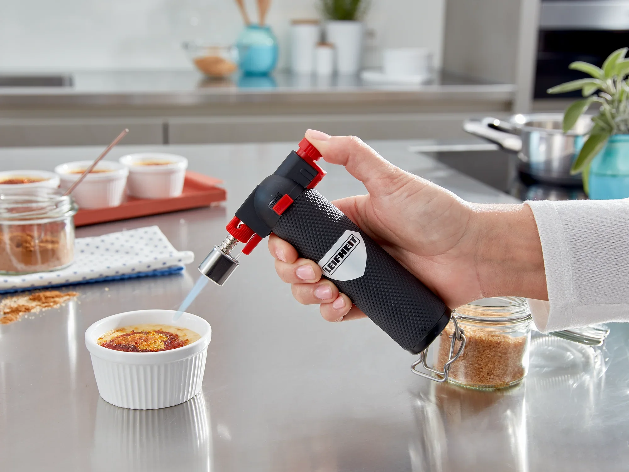 Ein Flambierer von Leifheit wird verwendet, um die Oberfläche eines Desserts in einer weißen Schale zu karamellisieren. Im Hintergrund sind weitere Desserts und Küchenutensilien zu sehen.