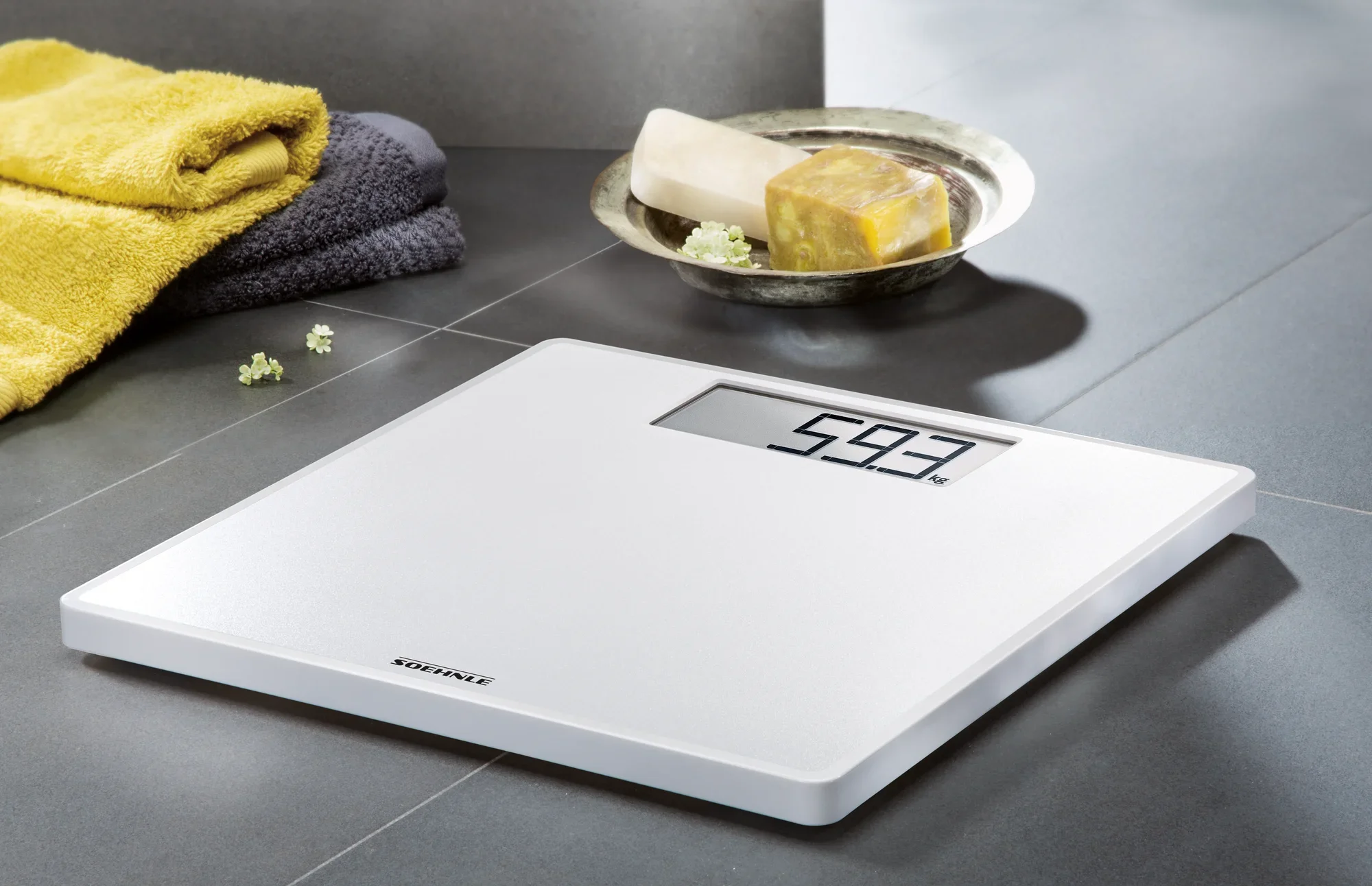 Cyfrowa waga łazienkowa Style Sense Save 100 na podłodze w łazience. Waga wyświetla 59,3 kg. W tle widoczne są ręczniki i kostki mydła.
