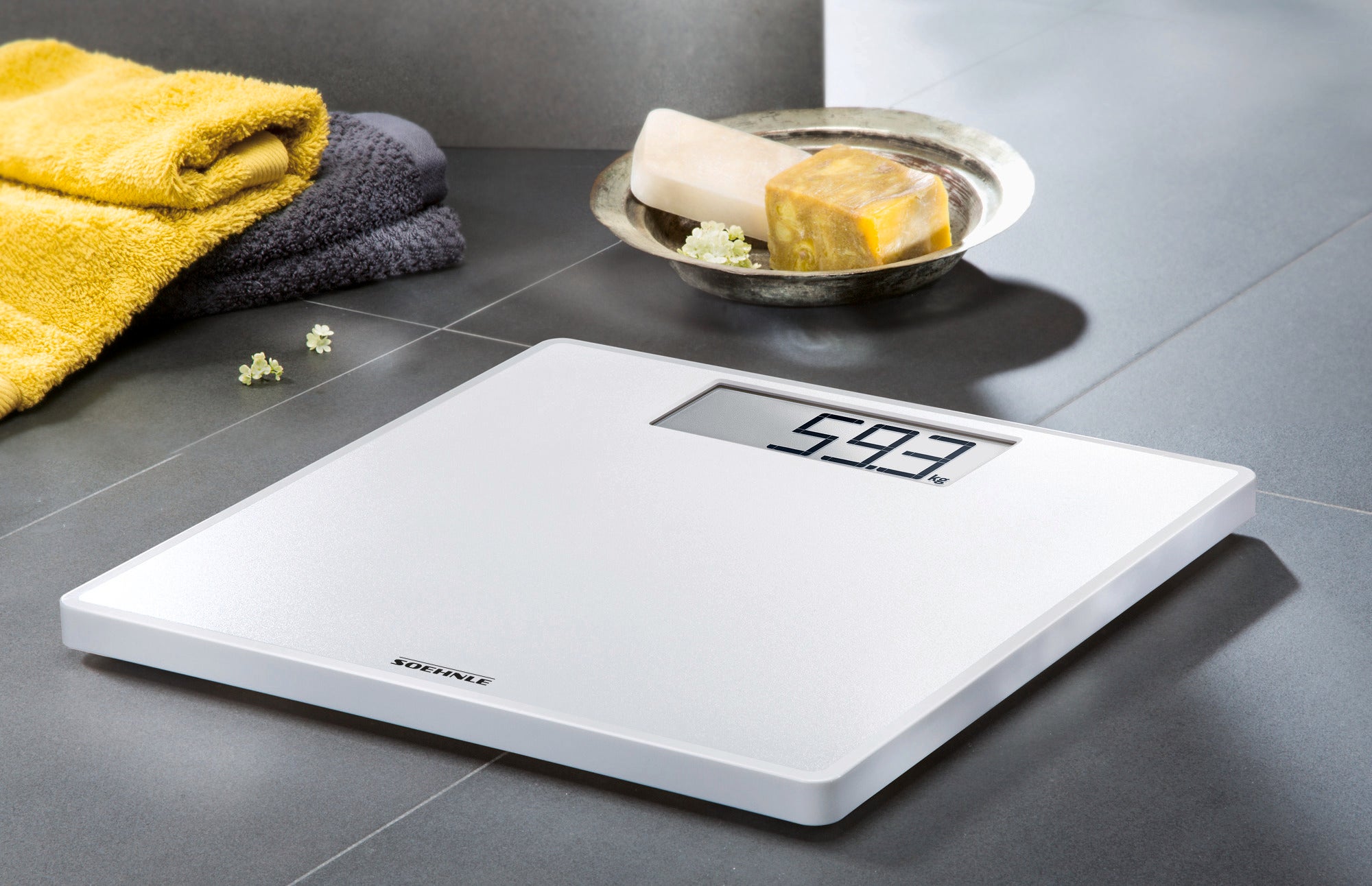 Personenwaage digital Style Sense Save 100 auf einem Badezimmerboden. Die Waage zeigt 59,3 kg an. Im Hintergrund sind Handtücher und Seifenstücke zu sehen.