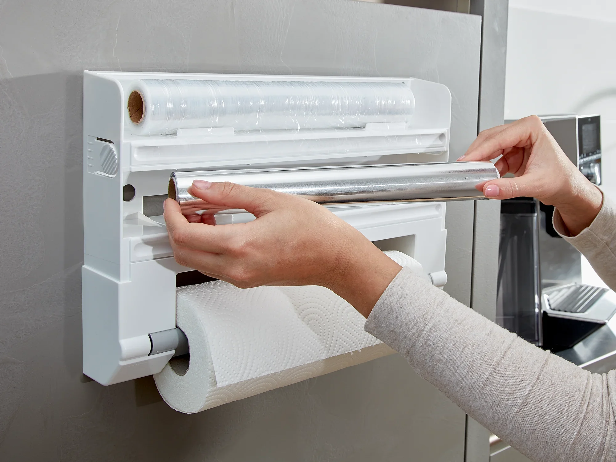 Support mural pour rouleaux Parat fixé à un mur de cuisine. Une personne change le papier aluminium. Le support offre de la place pour le papier aluminium et le papier essuie-tout, pratique et peu encombrant.