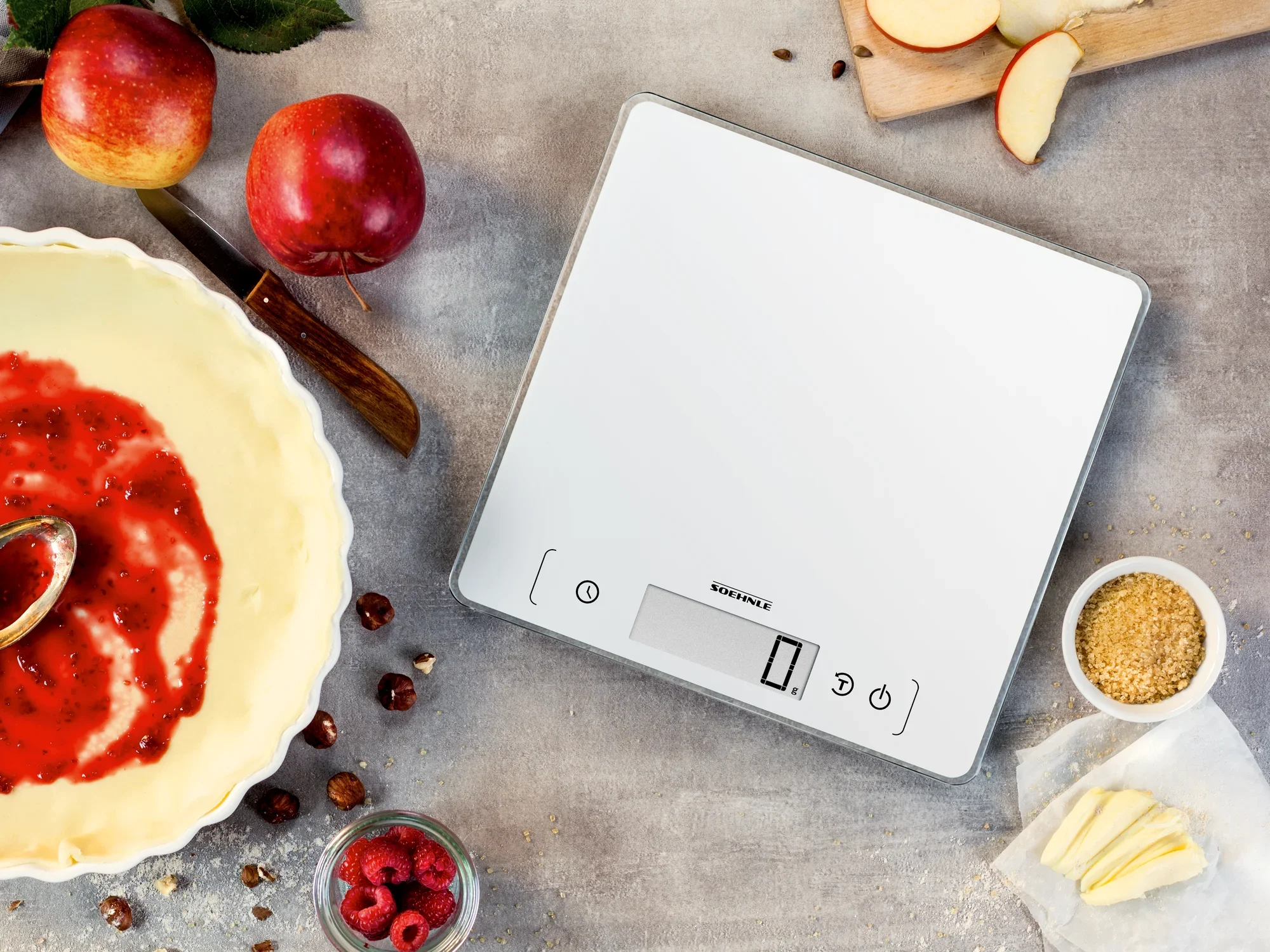 Digitale Küchenwaage Page Comfort 400 auf einer Küchenarbeitsplatte, umgeben von Äpfeln, Himbeeren, Nüssen und einer Schüssel mit rotem Teig. Ideal zum präzisen Abwiegen von Zutaten.