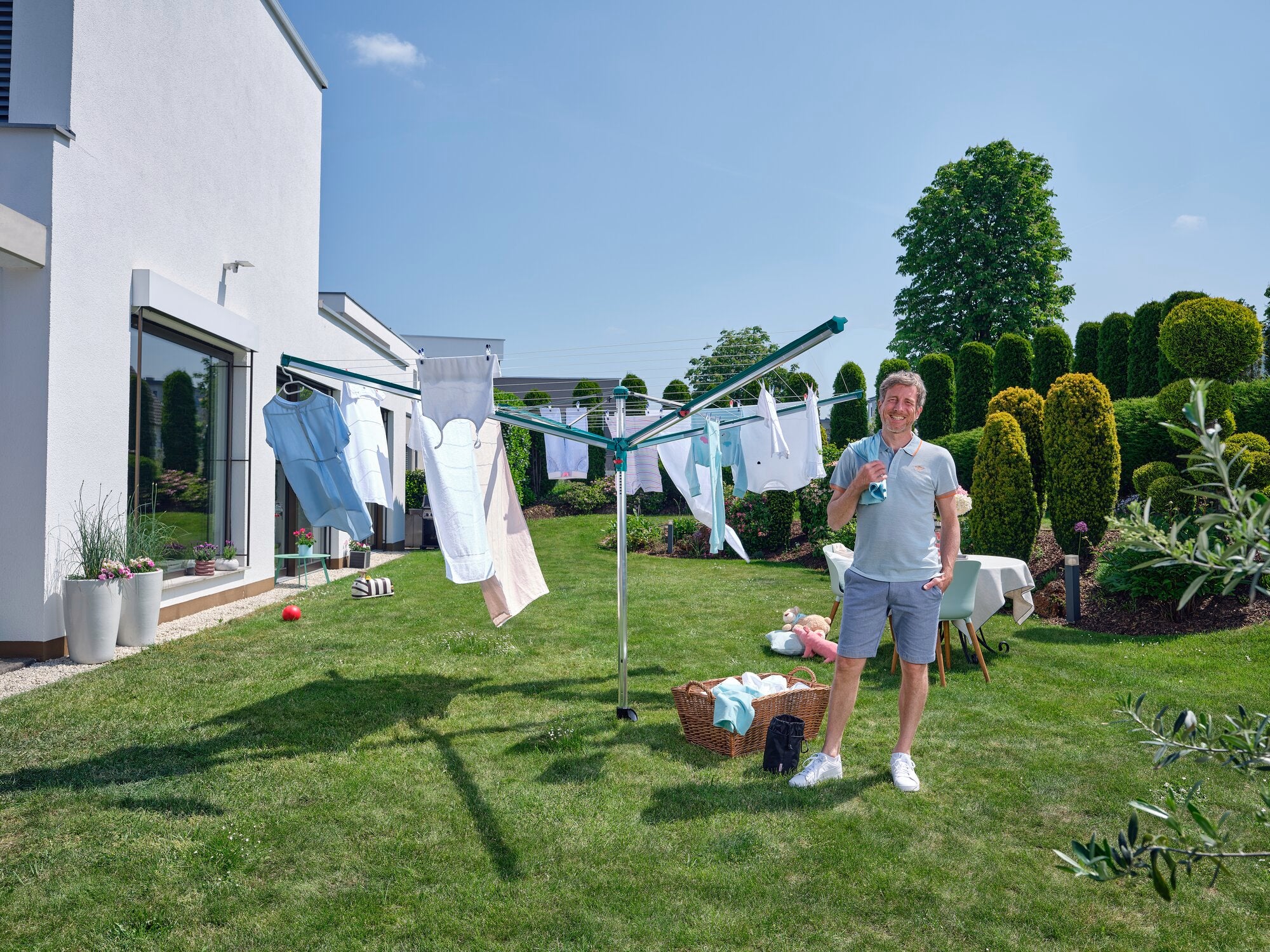 Ein Mann steht in einem sonnigen Garten neben einem Leifheit Linomatic 600 Deluxe Wäscheschirm. Frische Wäsche hängt zum Trocknen, während der Mann lächelt und ein Getränk hält.
