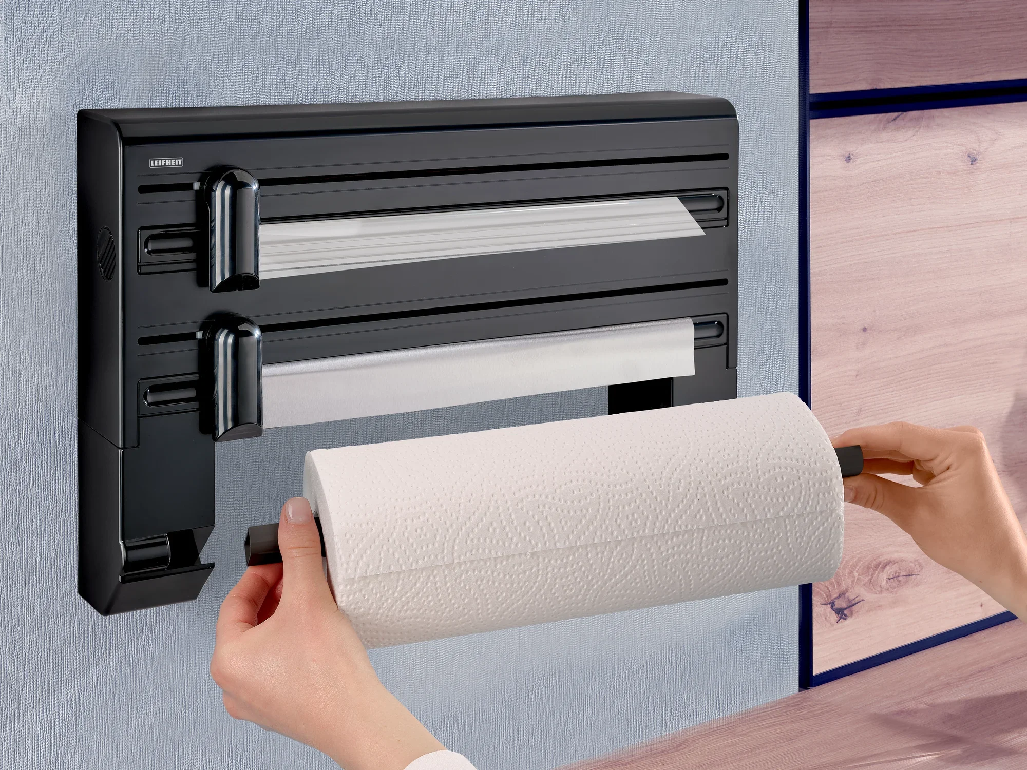 Der Leifheit Wandrollenhalter Parat Black ist an einer Wand montiert. Eine Person wechselt die Küchenrolle. Der Halter hat zwei weitere Fächer für Folien.