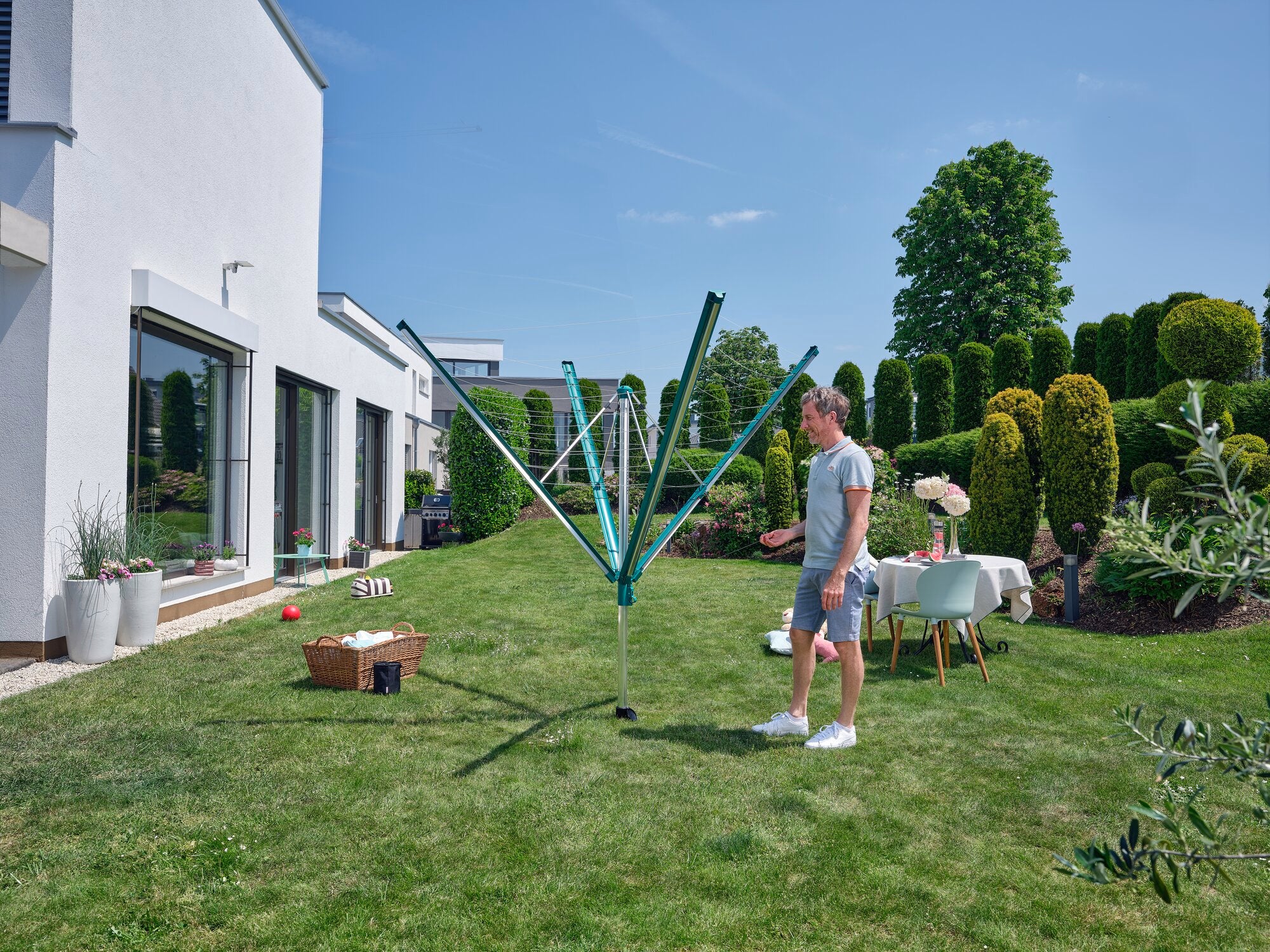 Ein Mann steht im Garten neben einem geöffneten Wäscheschirm Linomatic 600 Deluxe. Der Himmel ist klar, und im Hintergrund sind ein Haus und gepflegte Hecken zu sehen.