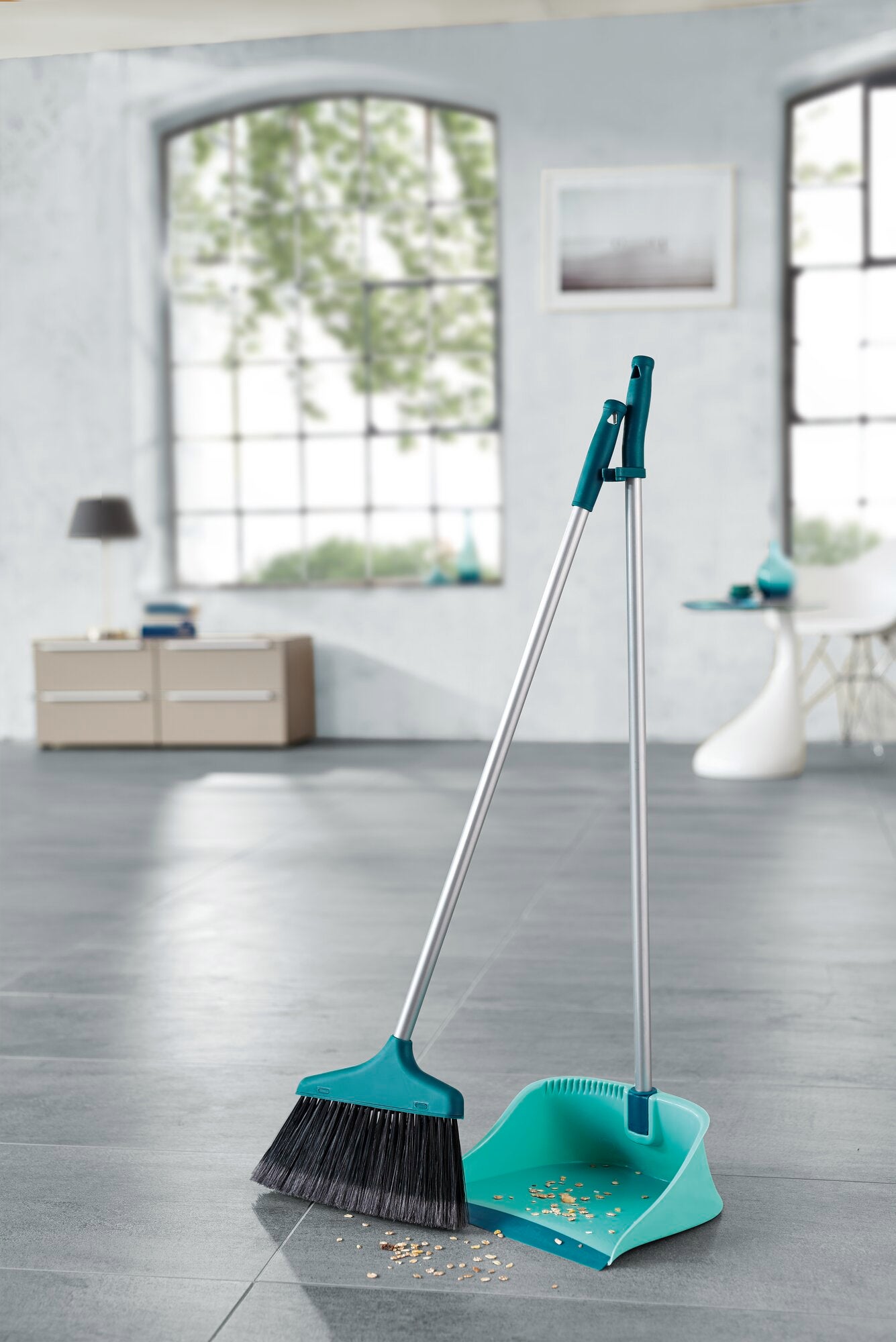 Set de limpieza Classic en un salón moderno. El set consta de una escoba azul y un recogedor, que se encuentran sobre un suelo gris. Al fondo se ven grandes ventanas y muebles.