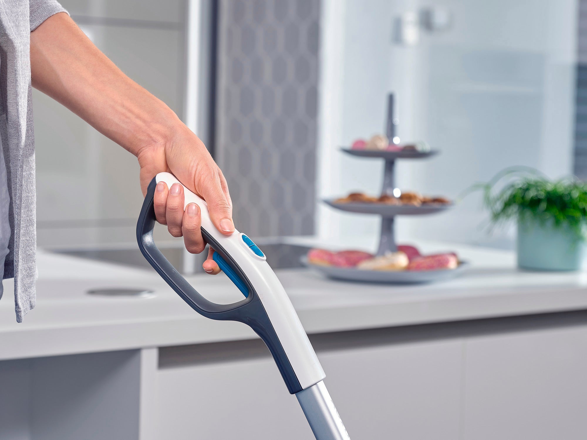 Eine Person hält den Griff des Regulus Aqua PowerVac, eines modernen Staubsaugers, in einer stilvollen Küche. Im Hintergrund sind ein Pflanzenarrangement und ein Serviertablett zu sehen.