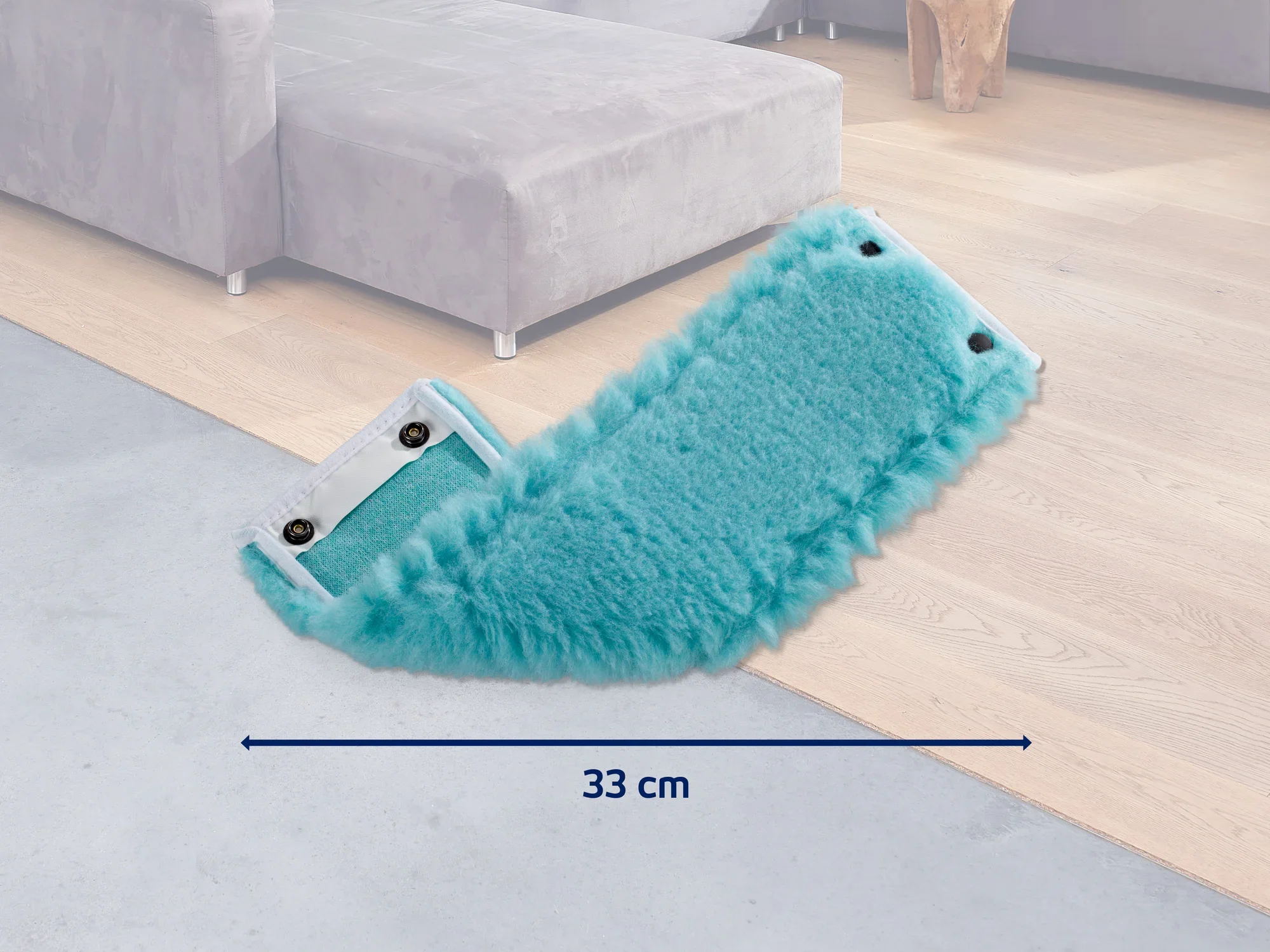 Een turkooizen mop met een breedte van 33 cm ligt op een lichtgekleurde houten vloer naast een grijze bank. Ideaal voor het grondig reinigen van vloeren.