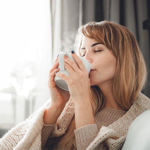 Eine Frau mit langen Haaren sitzt eingekuschelt in einer Decke und geniesst eine heisse Tasse Tee.