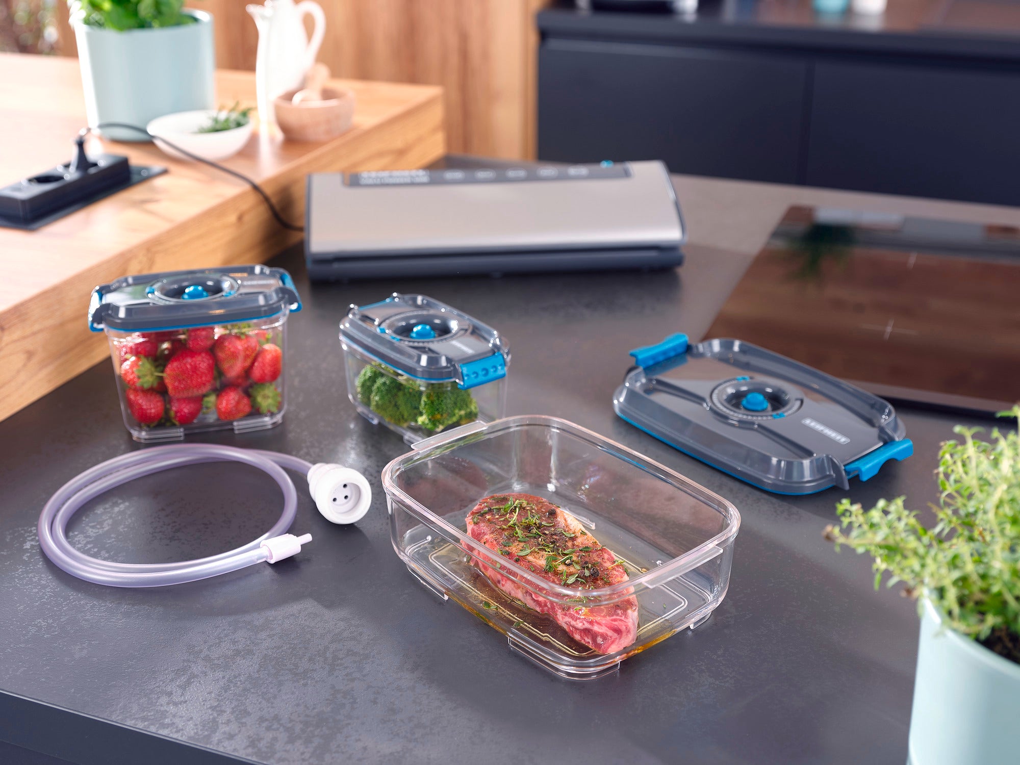 Vakuum-Frischhaltedosen-Set auf einer Küchenarbeitsplatte. Enthält Dosen mit Erdbeeren, Brokkoli und mariniertem Fleisch. Ein Vakuumiergerät und Schlauch sind ebenfalls sichtbar.
