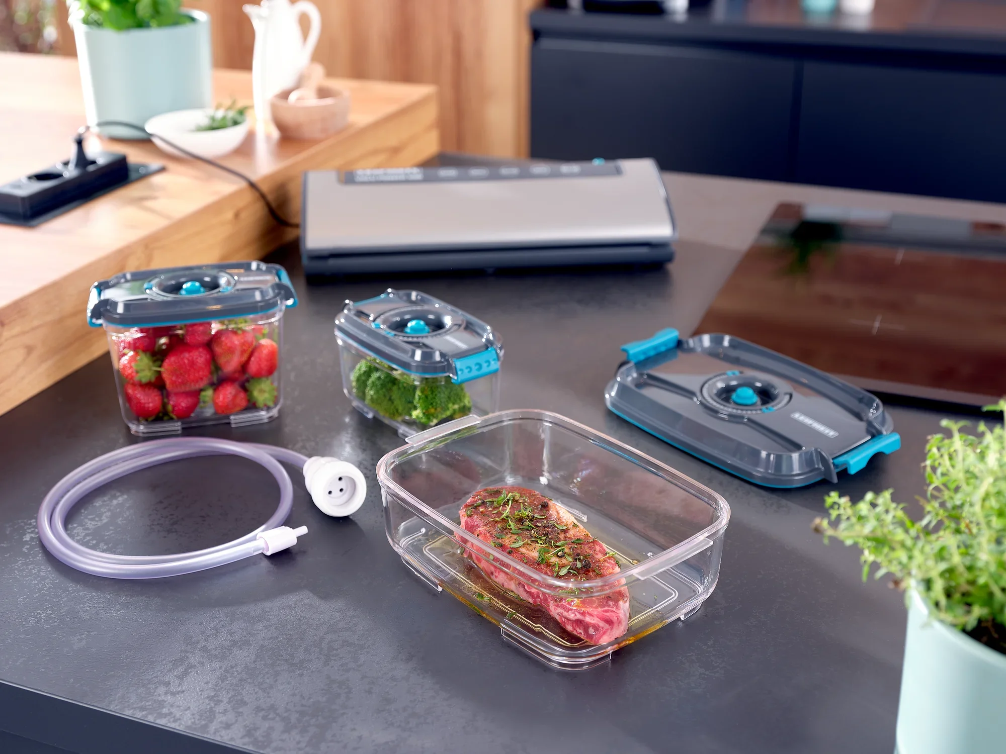 Zestaw próżniowych puszek do przechowywania żywności na blacie kuchennym. Zawiera puszki z truskawkami, brokułami i marynowanym mięsem. Widoczna jest również zgrzewarka próżniowa i wąż.