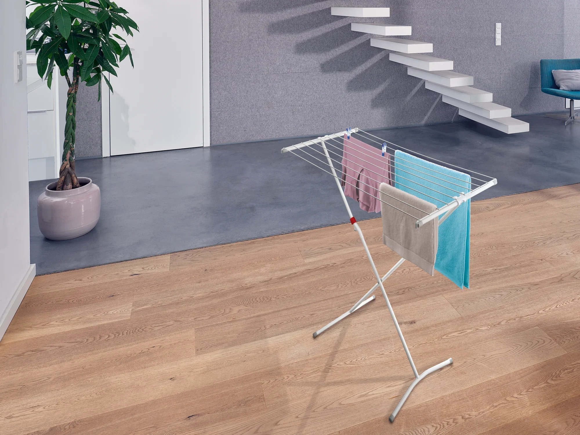 Wolnostojąca suszarka bębnowa Classic Easy 100 w nowoczesnym pomieszczeniu z drewnianą podłogą. Suszarka jest obwieszona kolorowymi ręcznikami i stoi obok rośliny i schodów.