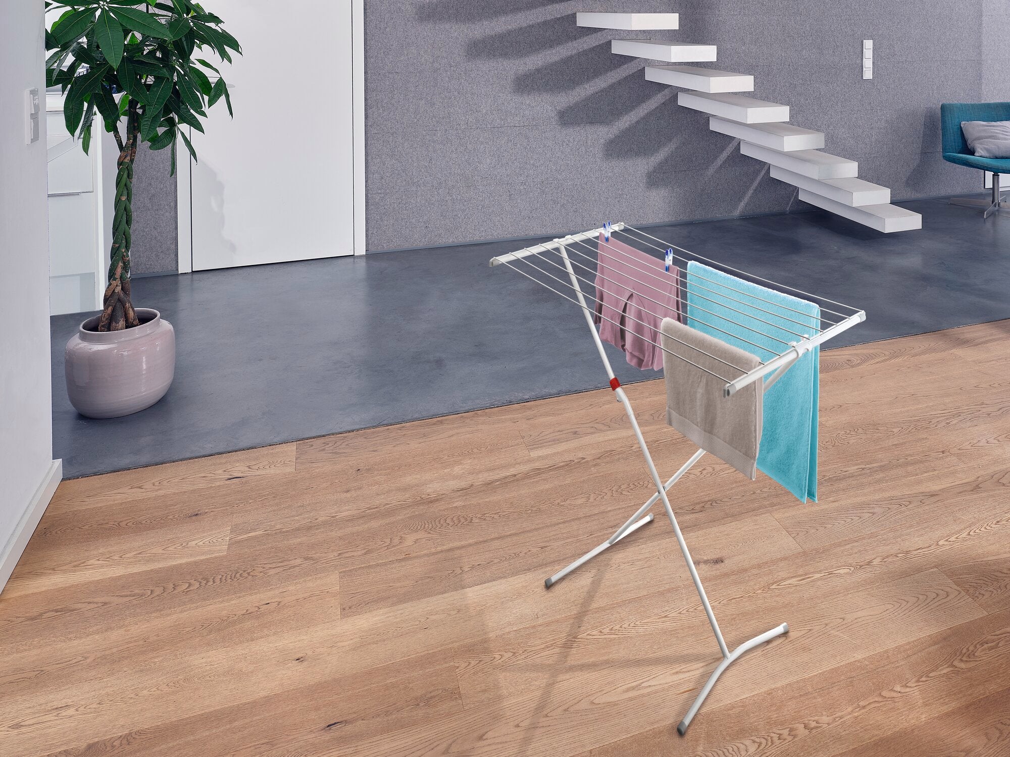 Wolnostojąca suszarka bębnowa Classic Easy 100 w nowoczesnym pomieszczeniu z drewnianą podłogą. Suszarka jest obwieszona kolorowymi ręcznikami i stoi obok rośliny i schodów.