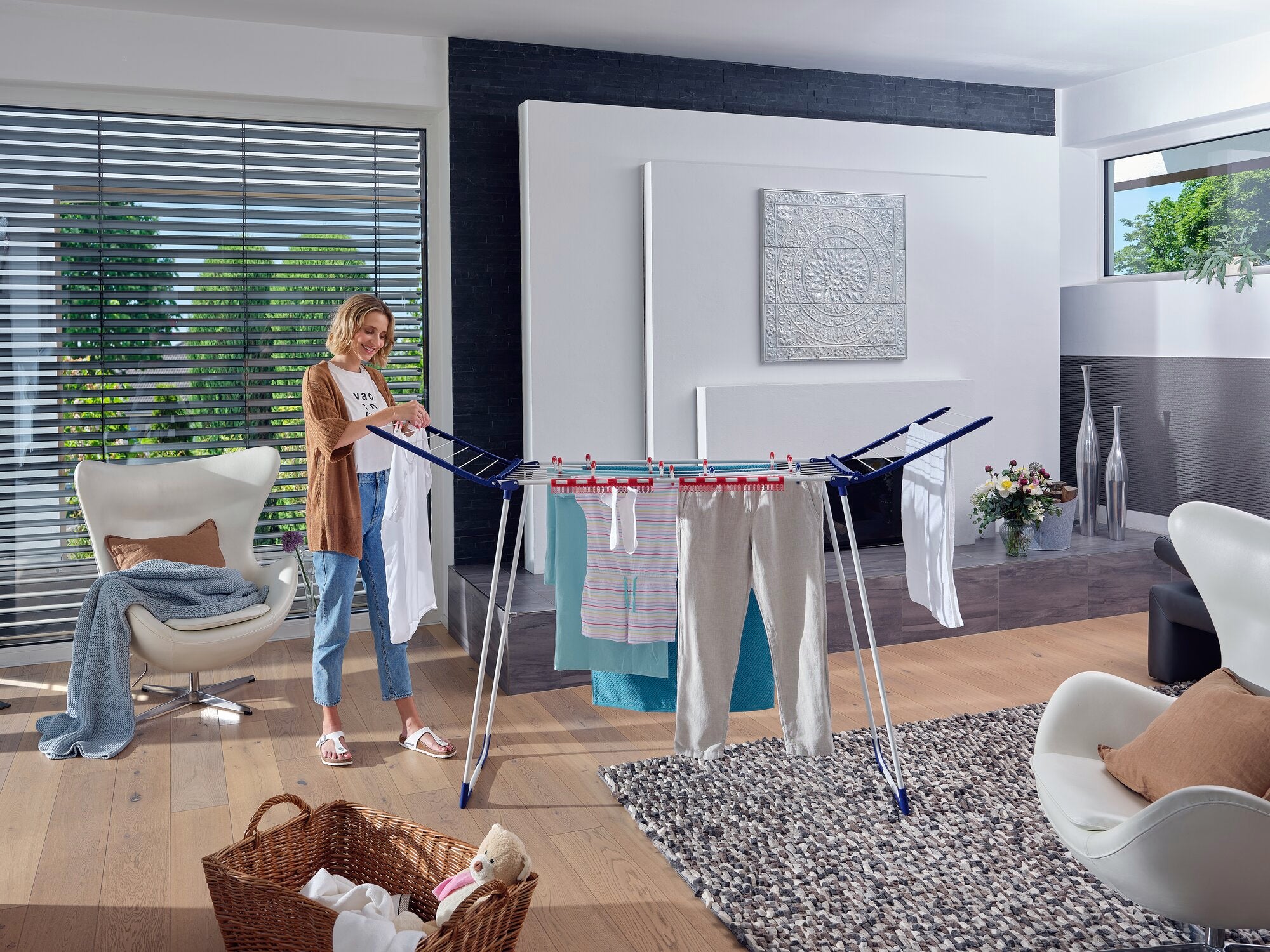 In einem modernen Wohnzimmer steht ein Standtrockner Pegasus 180 MAXX, auf dem Kleidung zum Trocknen aufgehängt ist. Eine Frau legt gerade ein weiteres Kleidungsstück darauf.