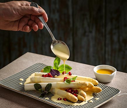Eine Hand giesst gelbe Sauce ueber weissen Spargel mit Granatapfelkernen und Basilikum auf einem gestreiften Teller neben einer Schale Sauce.