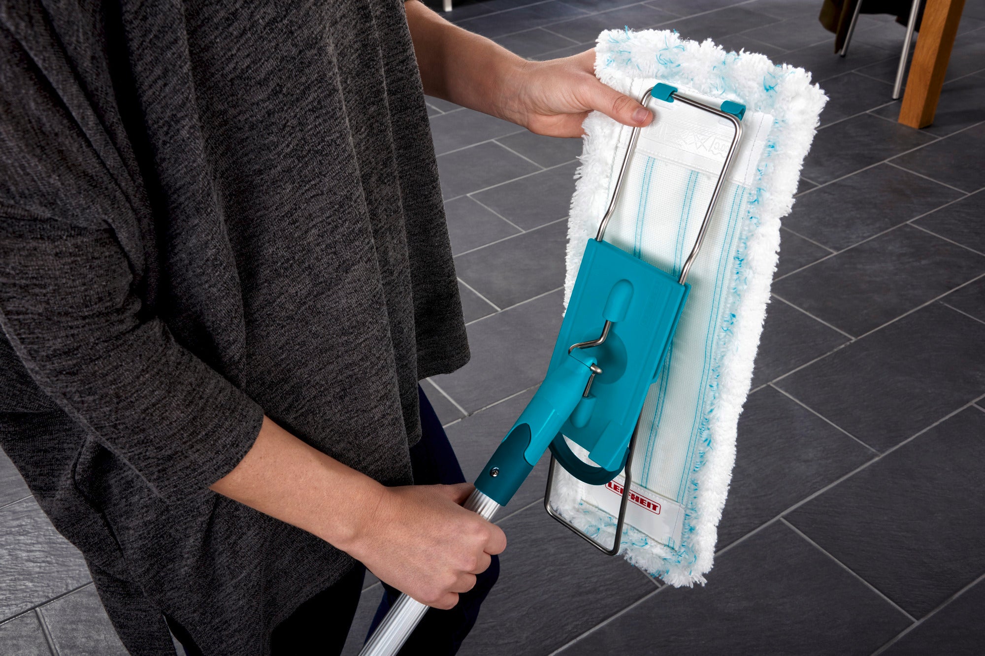 Eine Person benutzt den Bodenwischer Profi XL mit einem blauen Griff und einem weißen Mikrofaserbezug auf einem dunklen Fliesenboden. Der Wischer ist ideal für gründliche Reinigung und einfache Handhabung.