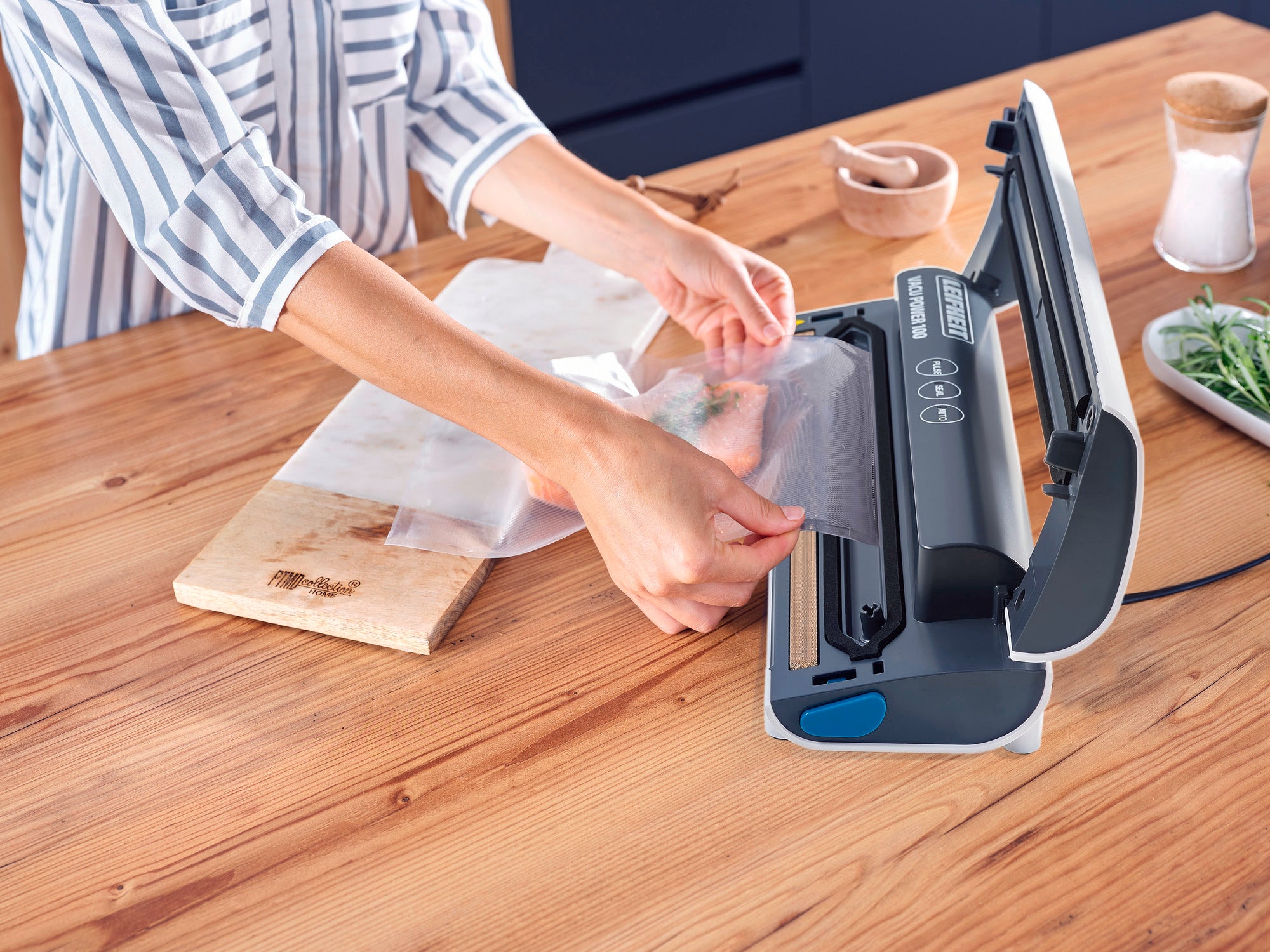 Eine Person verwendet das Vakuumiergerät Vacu Power 100, um Lebensmittel in einem Beutel zu versiegeln. Das Gerät steht auf einem Holztisch in einer modernen Küche.