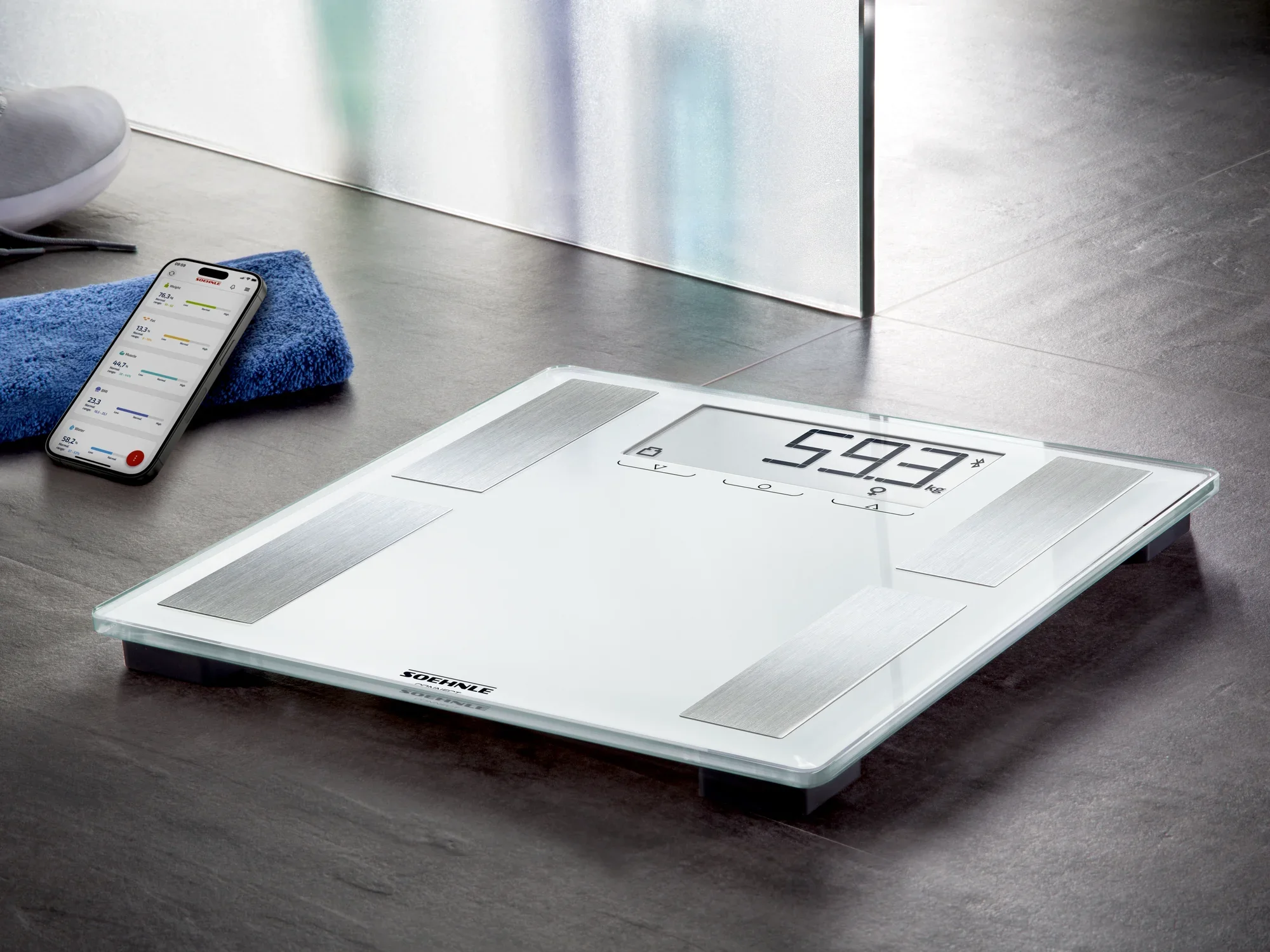 Shape Sense Connect 100 digitale badkamerweegschaal op een donkere vloer. Ernaast ligt een smartphone met een gezondheidsapp. De weegschaal geeft 59,3 kg aan.