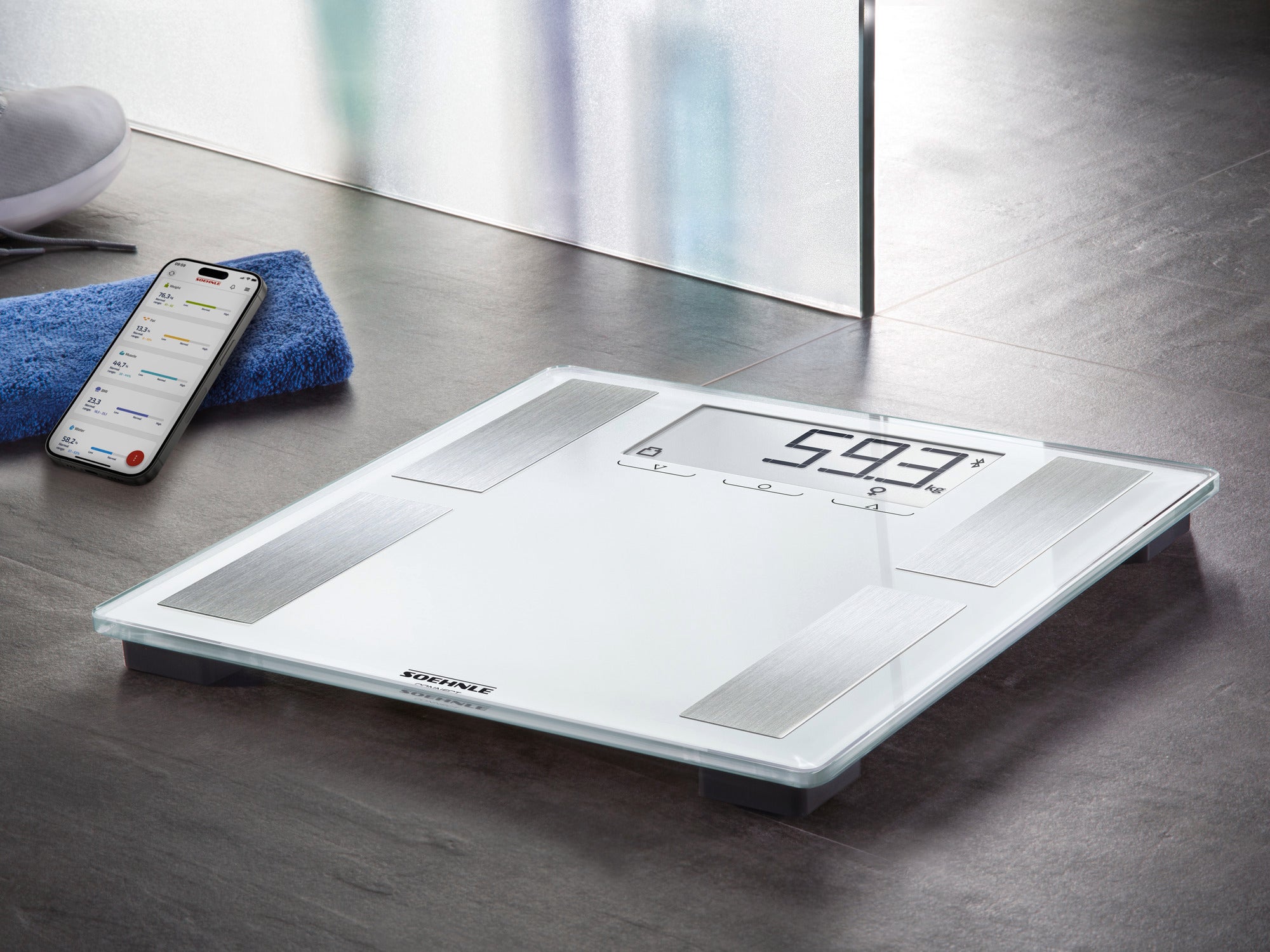 Digitale Personenwaage Shape Sense Connect 100 auf einem dunklen Boden. Ein Smartphone mit einer Gesundheits-App liegt daneben. Die Waage zeigt 59,3 kg an.