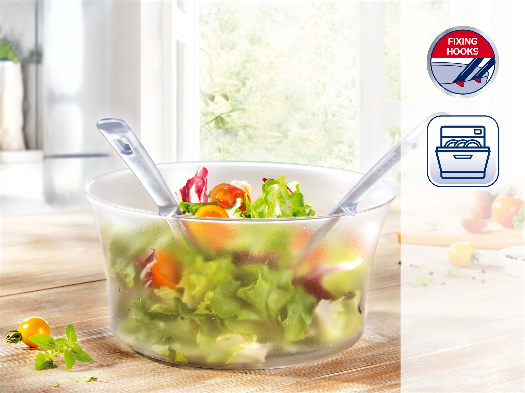 Leifheit Salatschleuder mit transparentem Behälter auf Holztisch, gefüllt mit grünem Salat und Tomaten; Deckel mit Kurbel, zwei Salatbestecke in der Schüssel, Symbol für aufhängbaren Deckel.