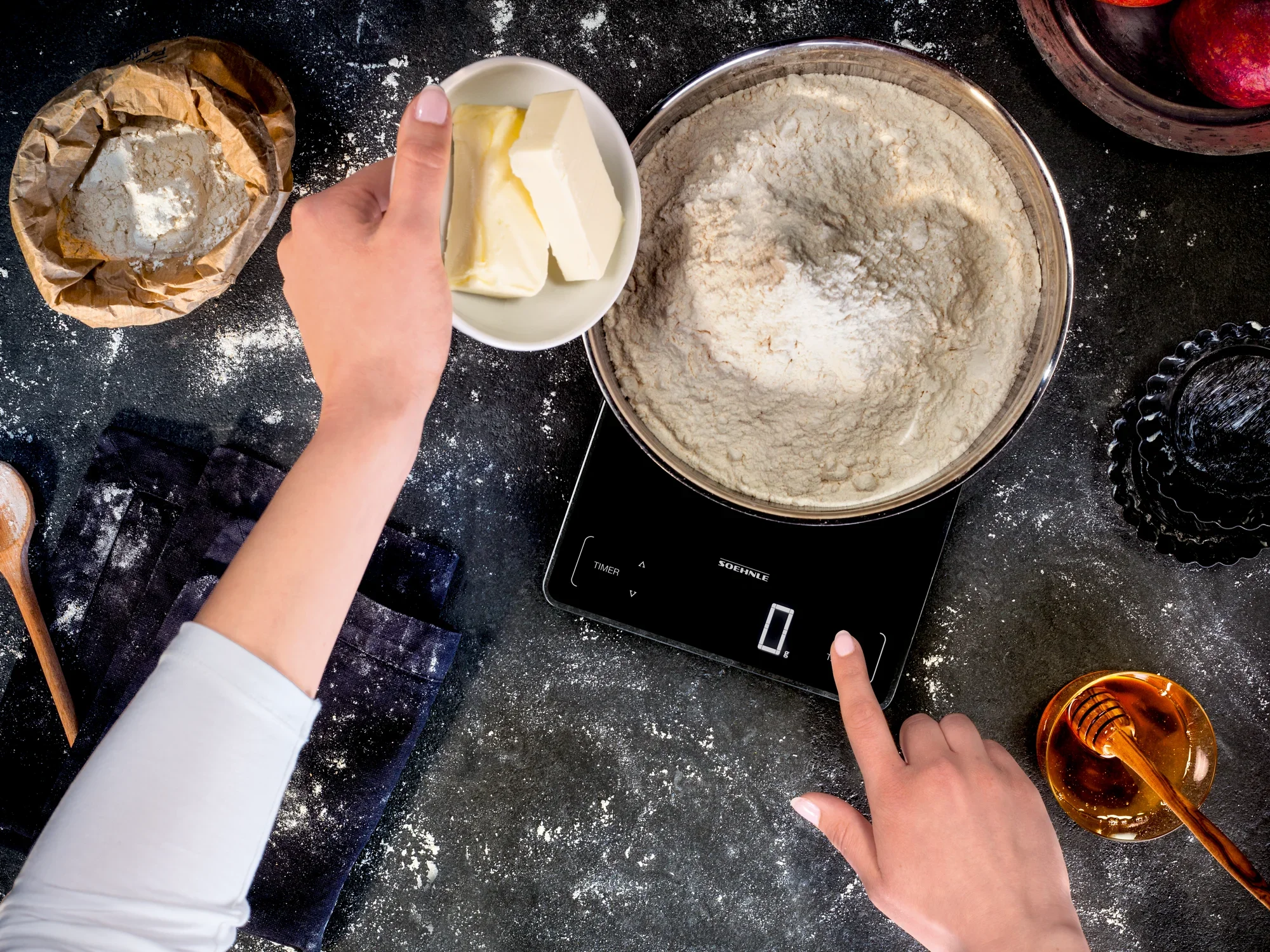 Een persoon weegt bloem op een digitale keukenweegschaal. Naast de weegschaal liggen ingrediënten zoals boter en suiker. De weegschaal geeft nauwkeurige metingen weer.
