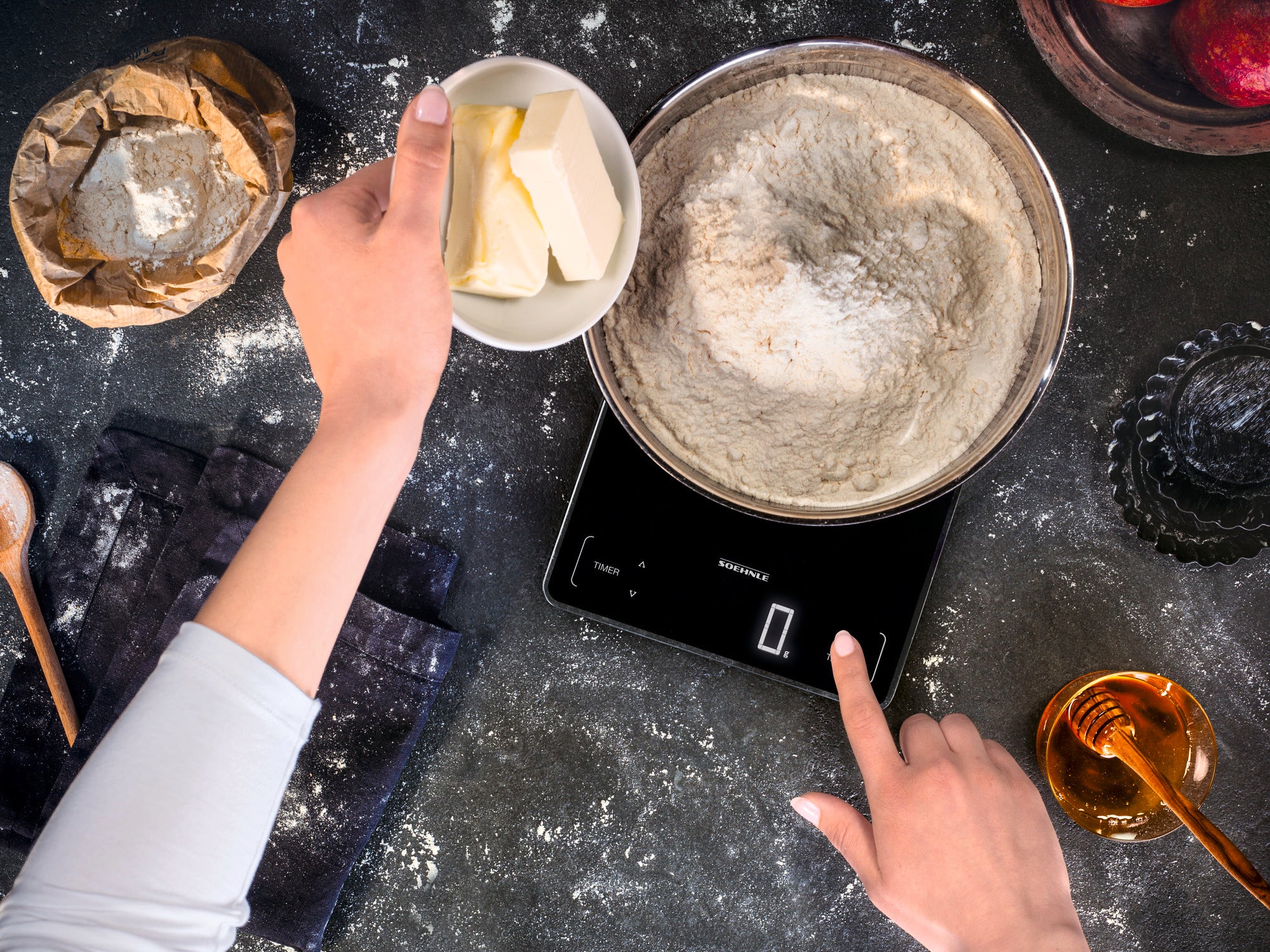 A person weighs flour on a digital kitchen scale. Ingredients such as butter and sugar are ready next to the scale. The scale displays precise measurements.