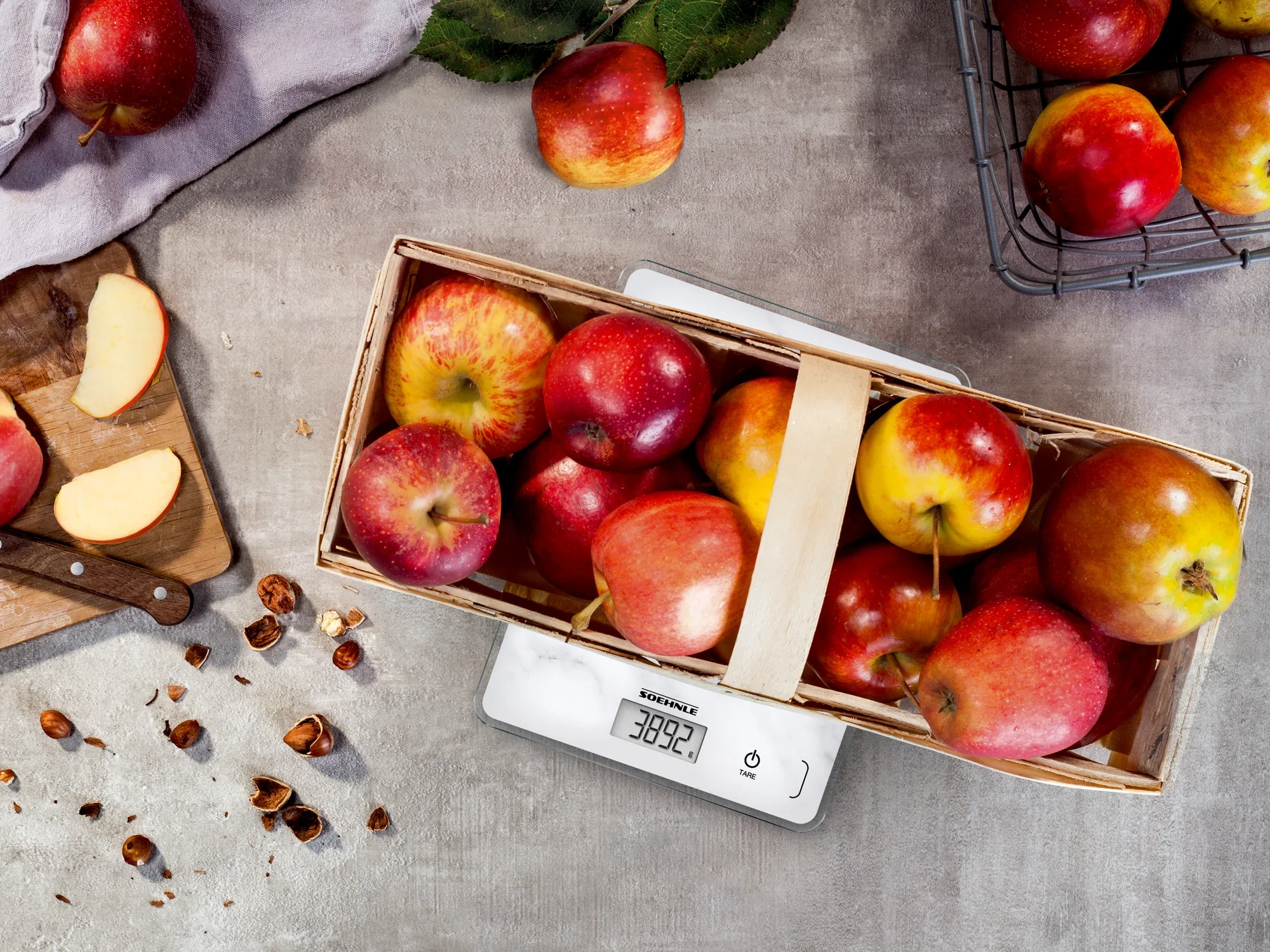A digital kitchen scale with a marble weighing surface displays the weight of a basket full of red apples. The scale is precise and stylishly placed in the kitchen.