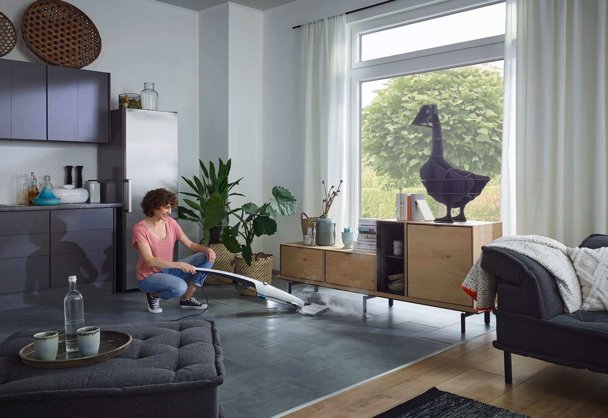 A person is using the CleanTenso Power steam cleaner on a grey tiled floor in a modern living room. Large windows let in plenty of daylight, while plants and furniture decorate the room.